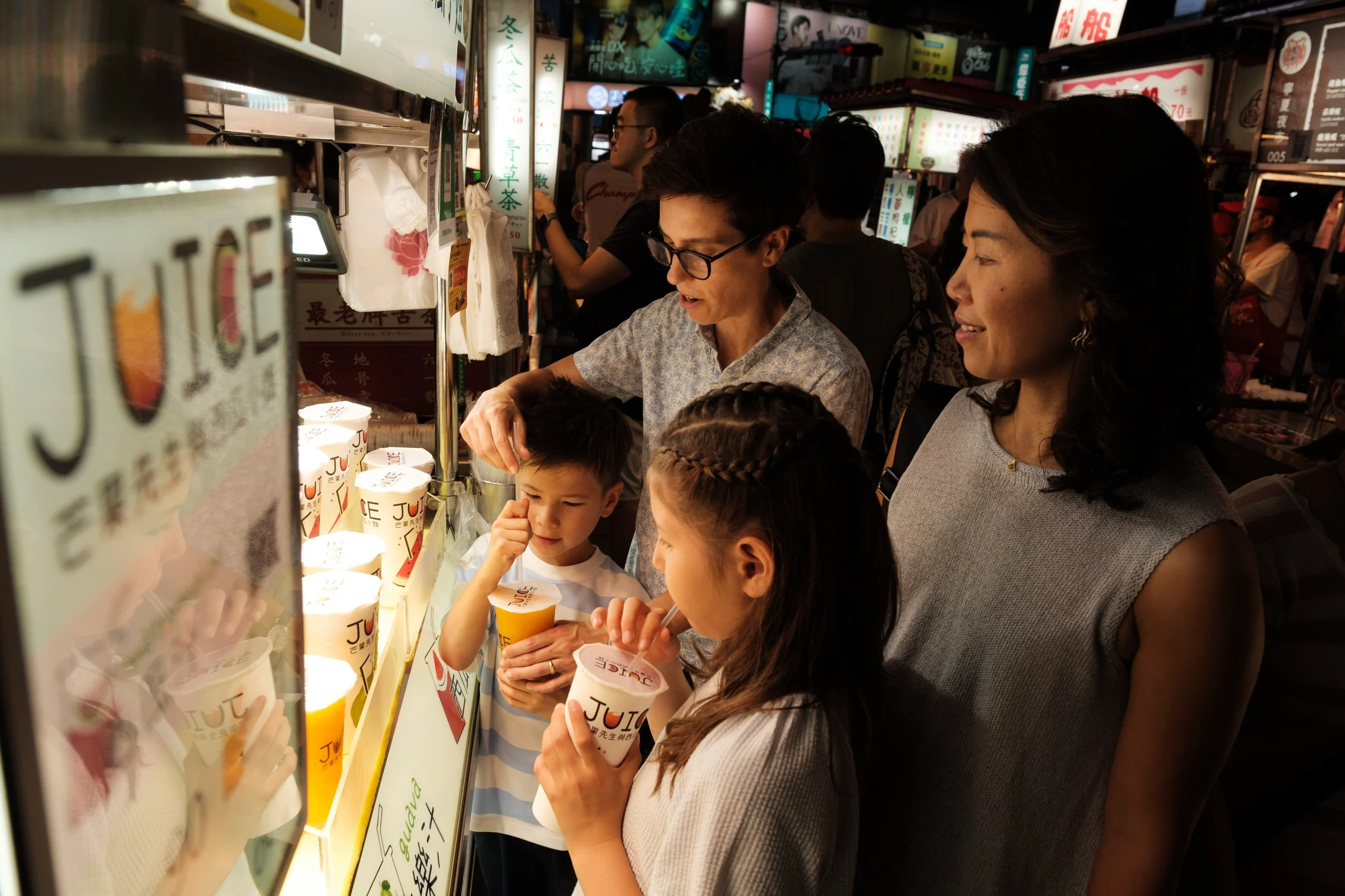 Two moms and their two kids crowd around a juice stall at Ningxia Night Market, the kids holding fresh-squeezed cups while one mom reaches to add a straw. Taipei family photography.