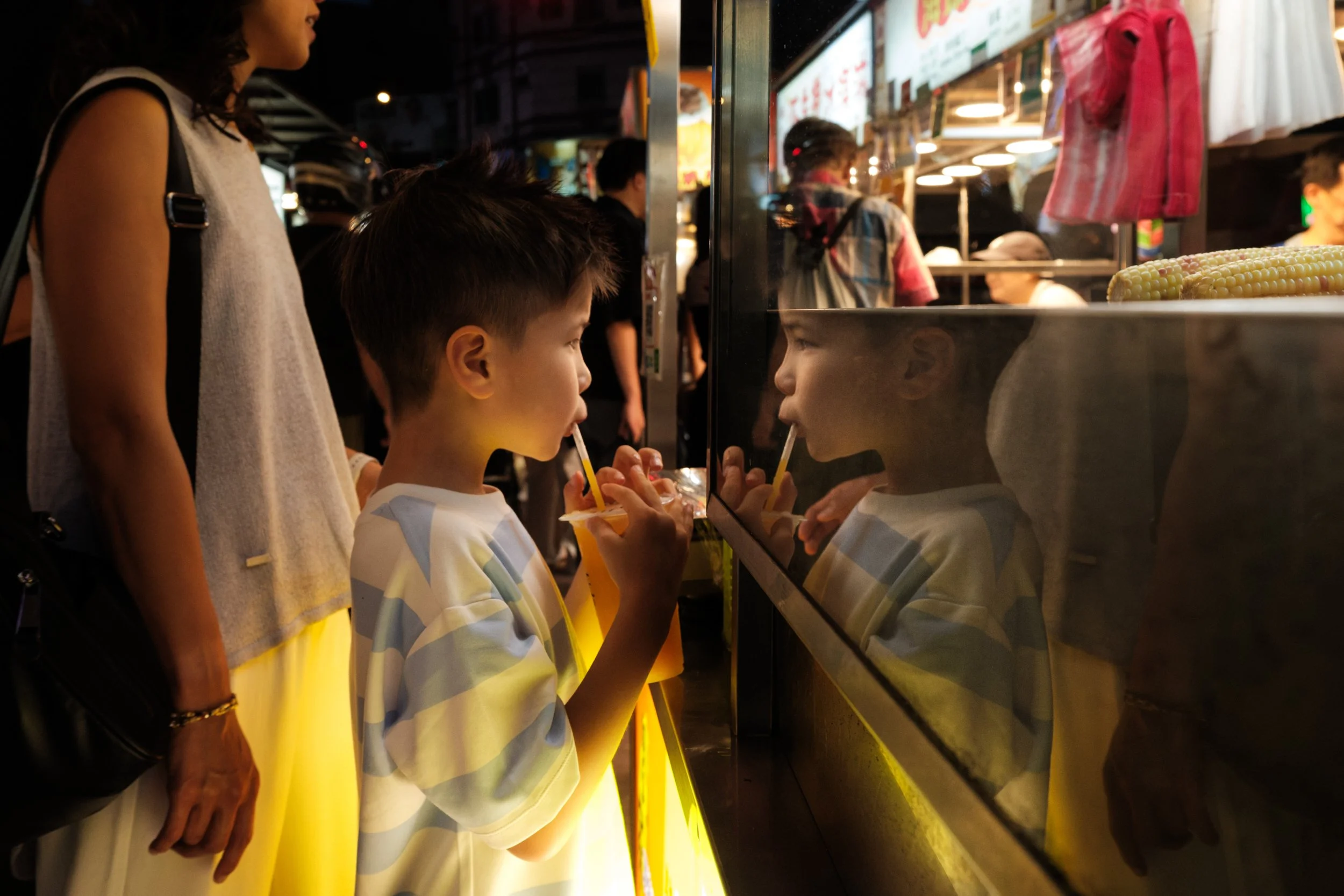 A boy sips juice through a straw at Ningxia Night Market, his face reflected in the glass of a lit stall beside him, his mom visible behind him. Taipei documentary family photography.