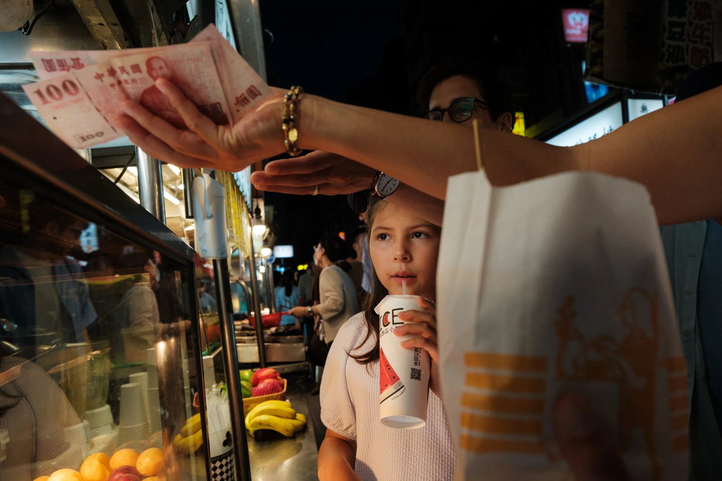 A girl sips juice at Ningxia Night Market while her mom pays overhead with NT$100 bills, the busy market stalls blurred behind them. Taipei outdoor family photoshoot.