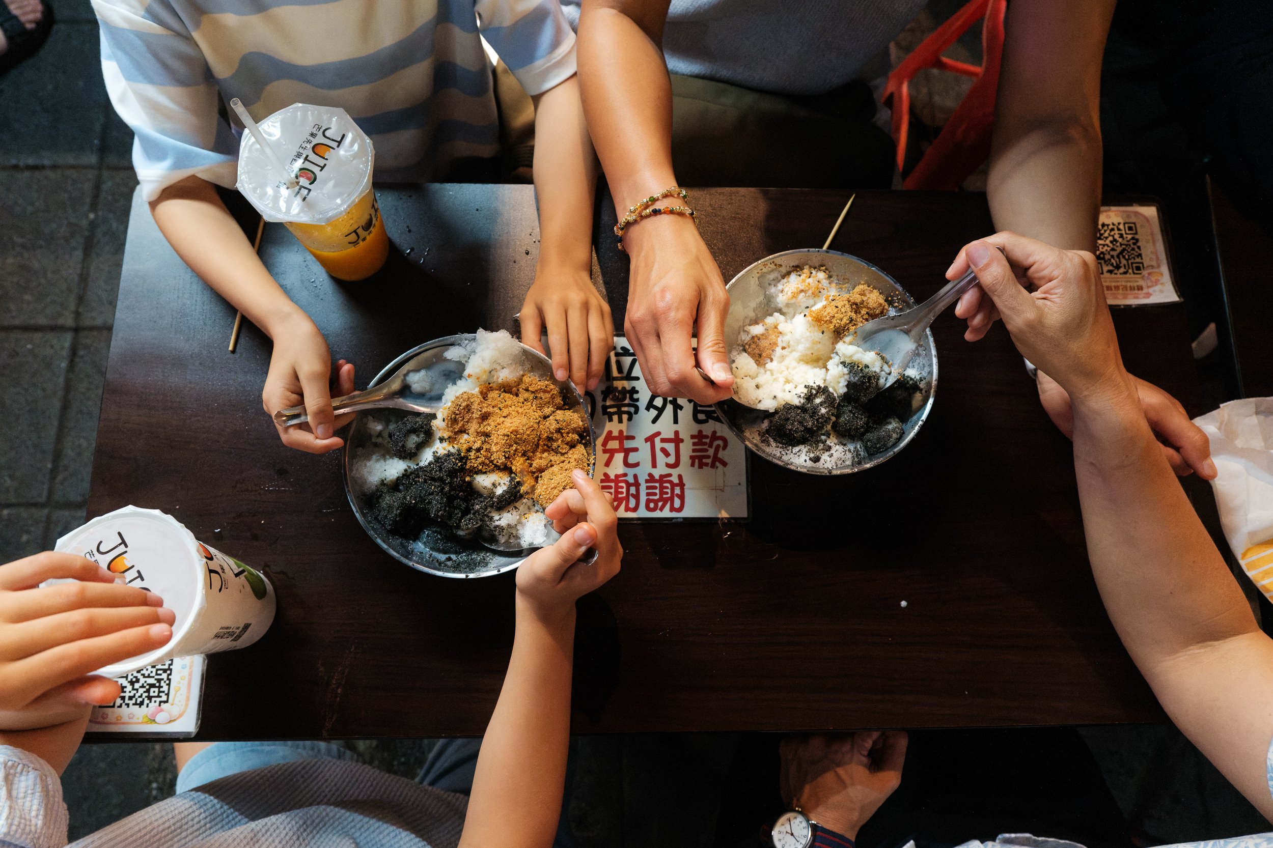 Overhead view of four pairs of hands sharing two bowls of mochi shaved ice at Ningxia Night Market, juice cups on the table beside them. Taipei family photography.