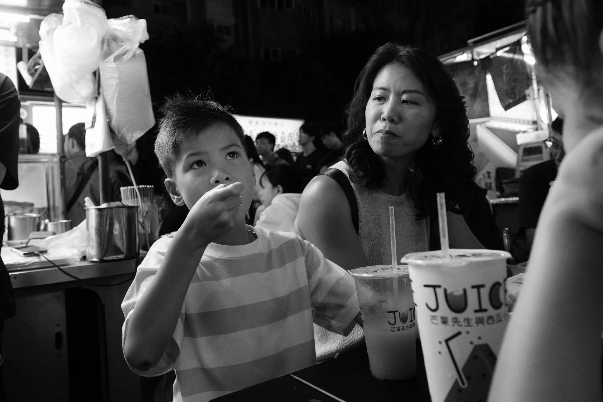 A boy and his mom sit side by side at Ningxia Night Market, both quiet, drinks in hand, watching the crowd. Black and white. Taipei documentary family photography.
