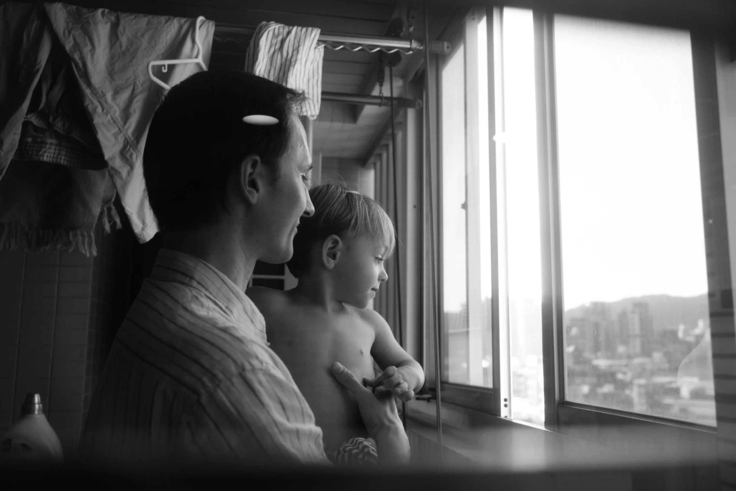 Taipei documentary family photography, Tienmu apartment | Father holds his shirtless toddler at a window overlooking the Taipei skyline