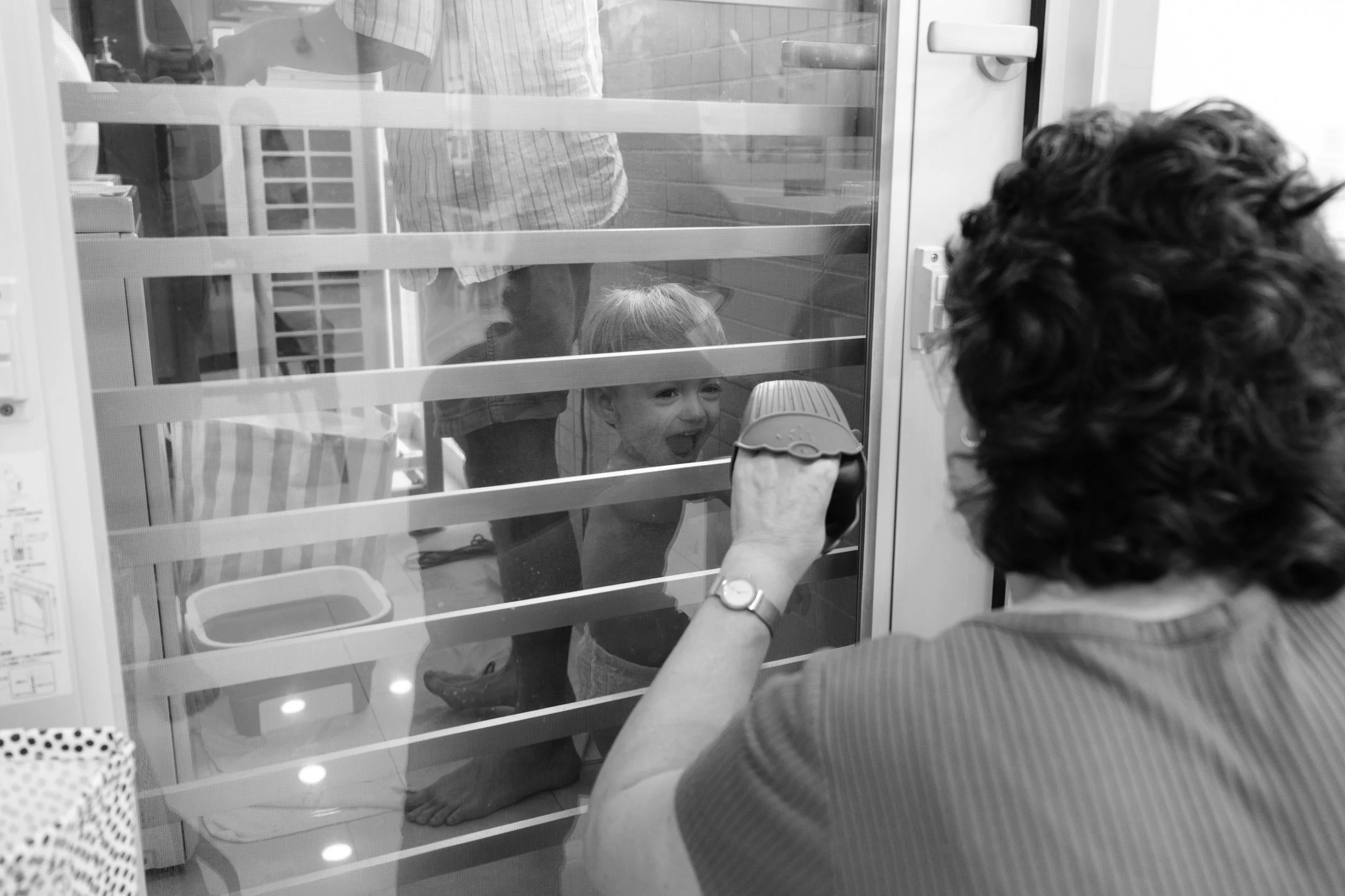 Taipei documentary family photography, Tienmu home | Toddler laughs through a balcony door screen as while his grandmother makes a silly face from the other side.