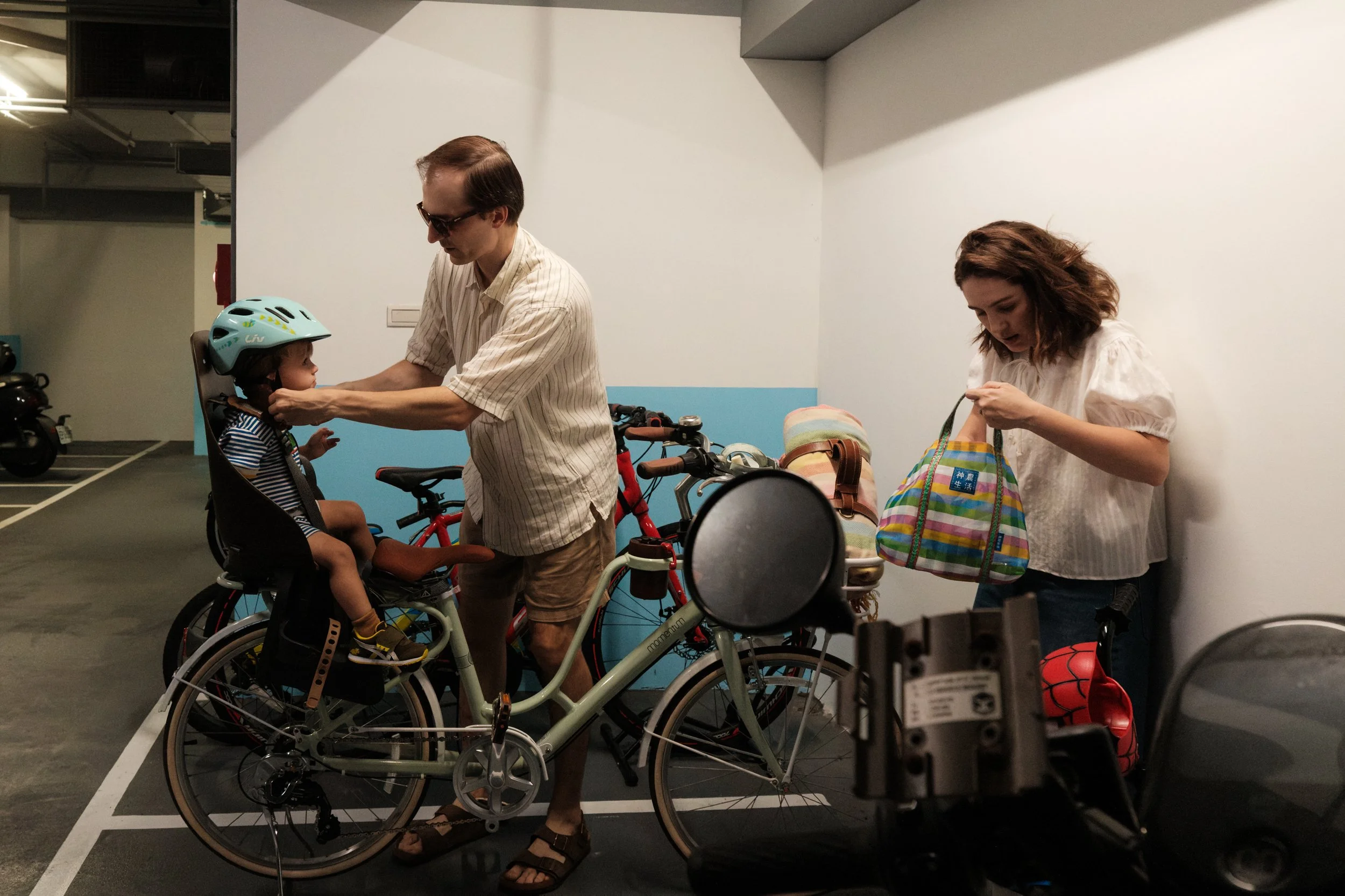 Taipei documentary family photography, Tienmu | Father buckles a toddler's helmet in a parking garage as the family prepares to bike out