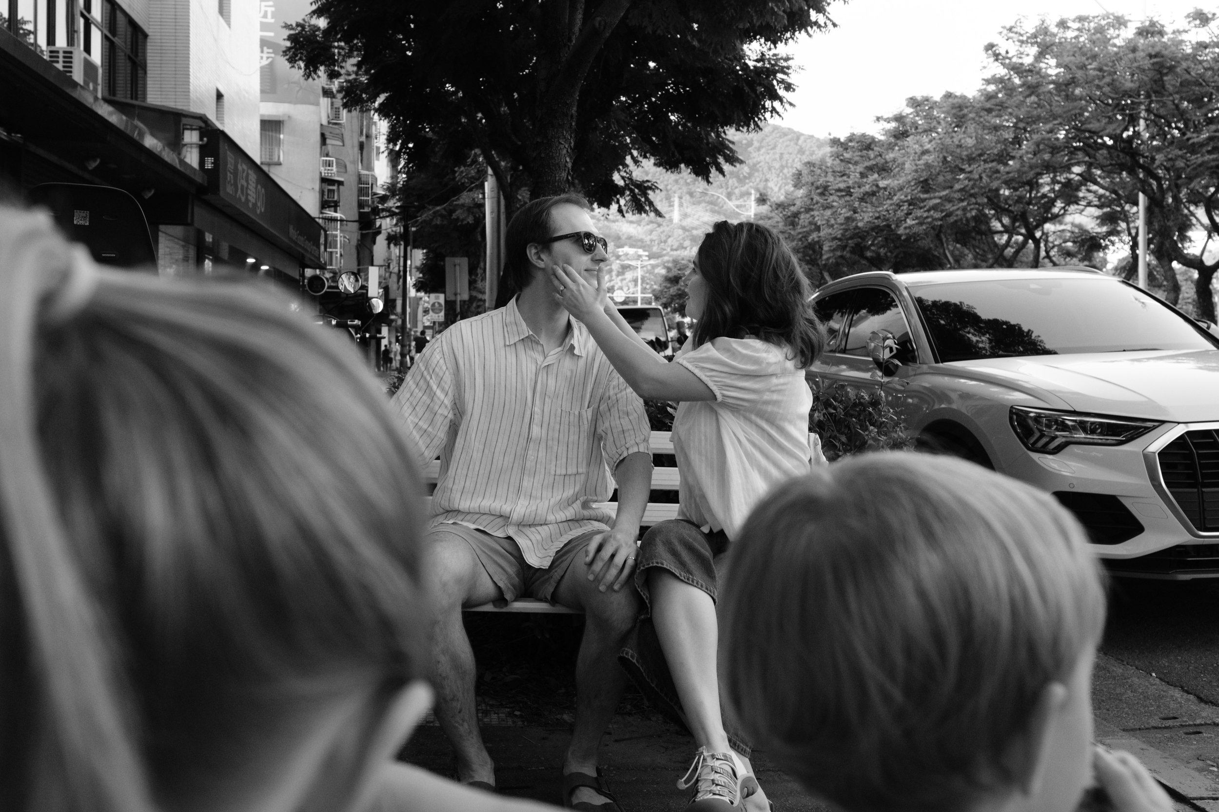 Taipei documentary family photography, Tienmu street | Wife cups her husband's face on a bench beneath a tree, framed by their children in the foreground