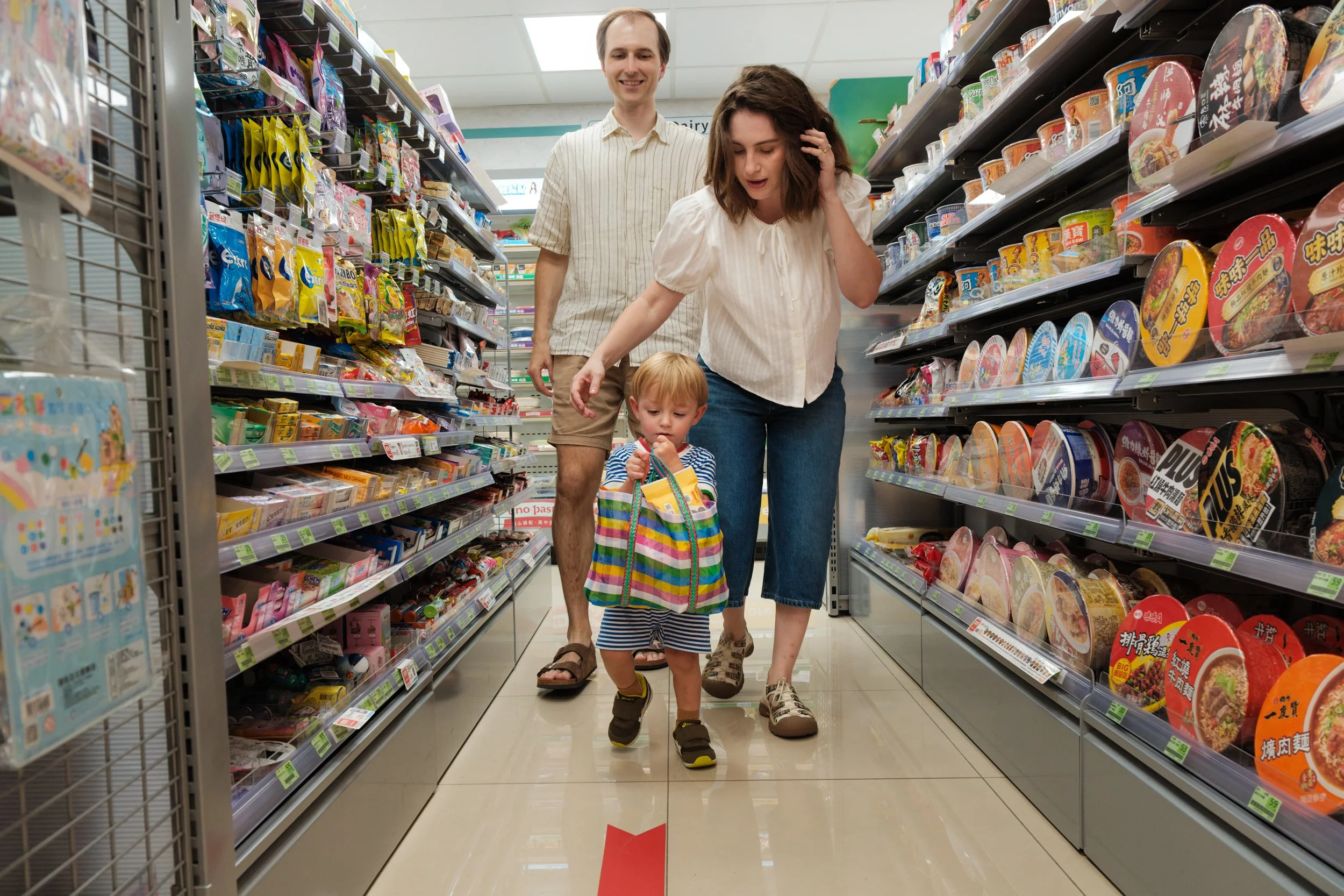 Taipei documentary family photography, Tienmu convenience store | Family walks together down a snack aisle, toddler carrying a striped market bag