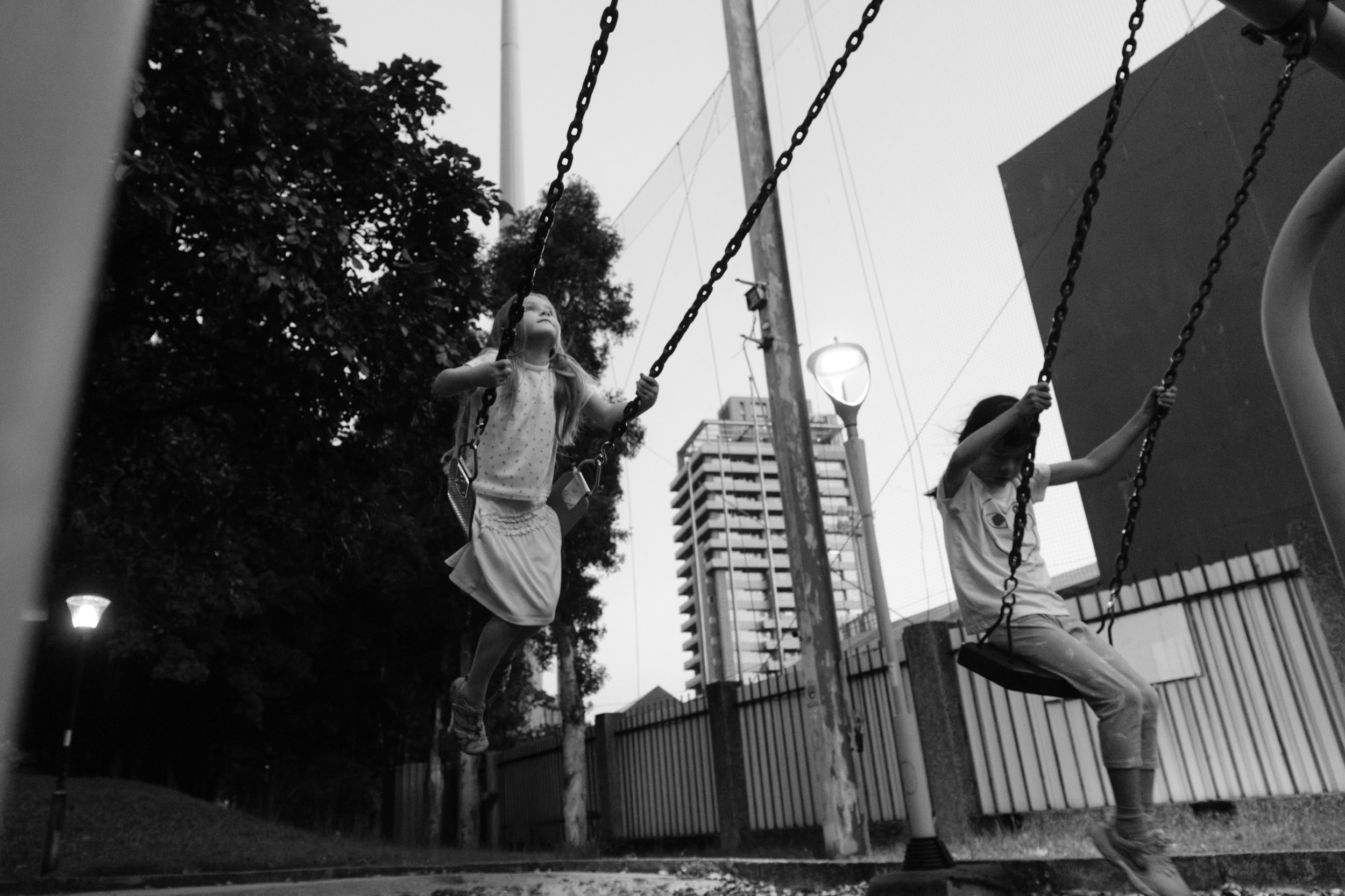 Taipei documentary family photography, Tienmu Sports Park | Two girls swing side by side at dusk with the Taipei skyline behind them
