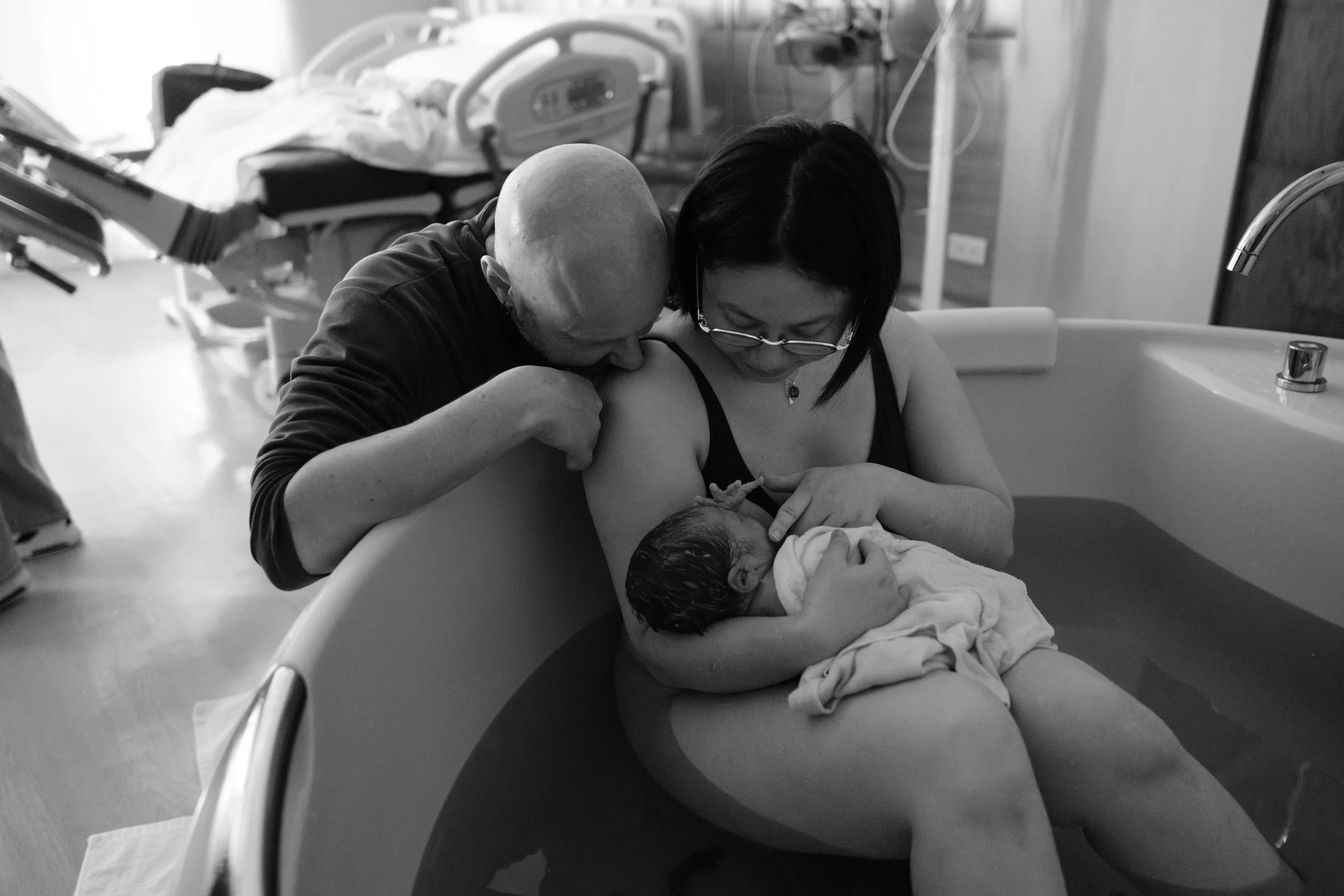 Birth photography at Dianthus, Taipei — father embracing mother and newborn in the water birth tub just after delivery.