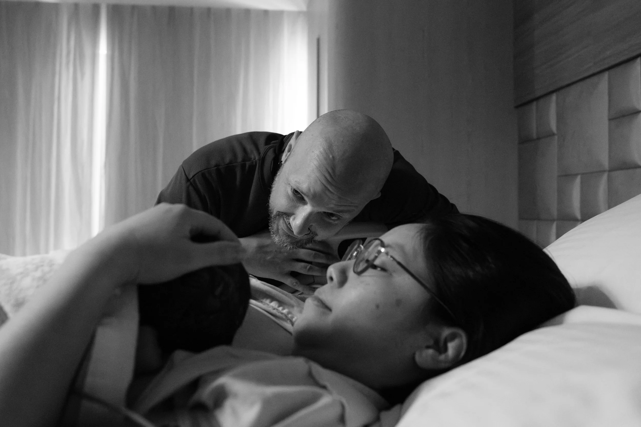 Taipei birth photography — father leaning close to mother's face as she rests in her Dianthus birth suite after delivery.