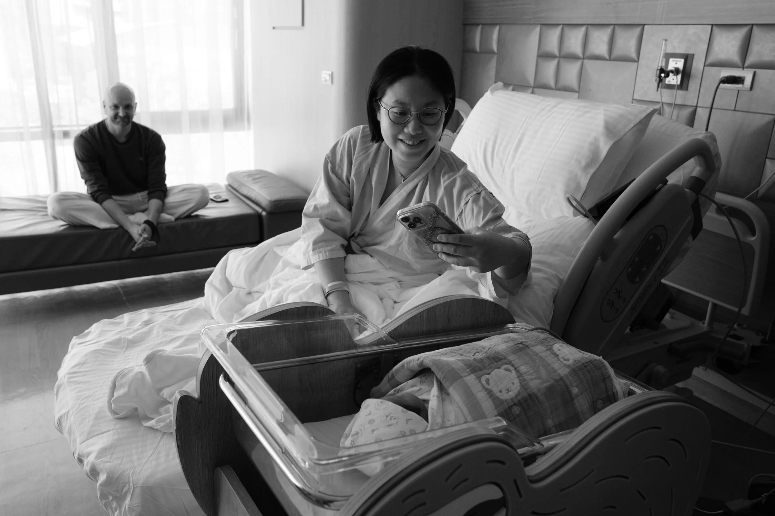Taipei birth photography at Dianthus — mother smiling at her baby in a bassinet in her postpartum room, taking a picture with her phone.