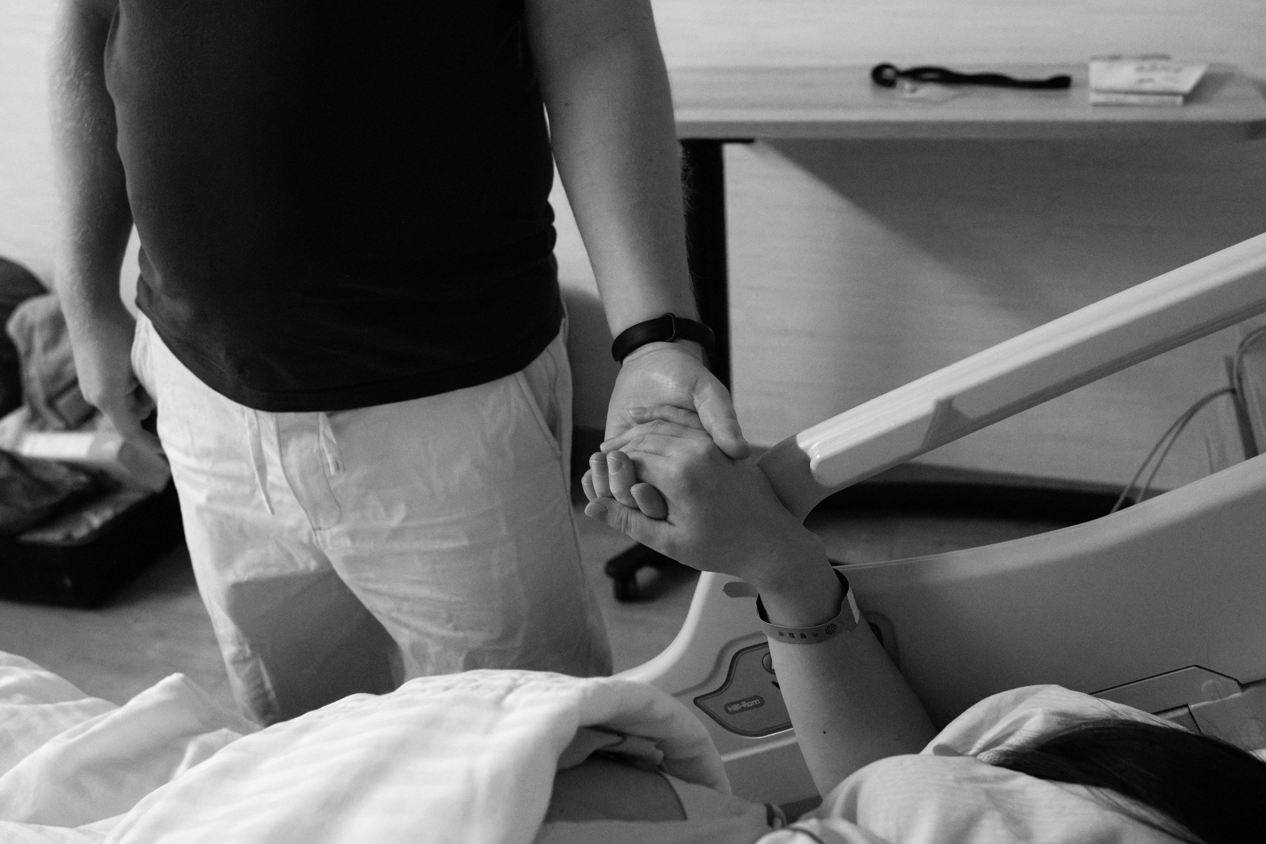 Birth photography at Dianthus Taipei — partner holds mother's hand beside the hospital bed before c-section surgery, black and white.