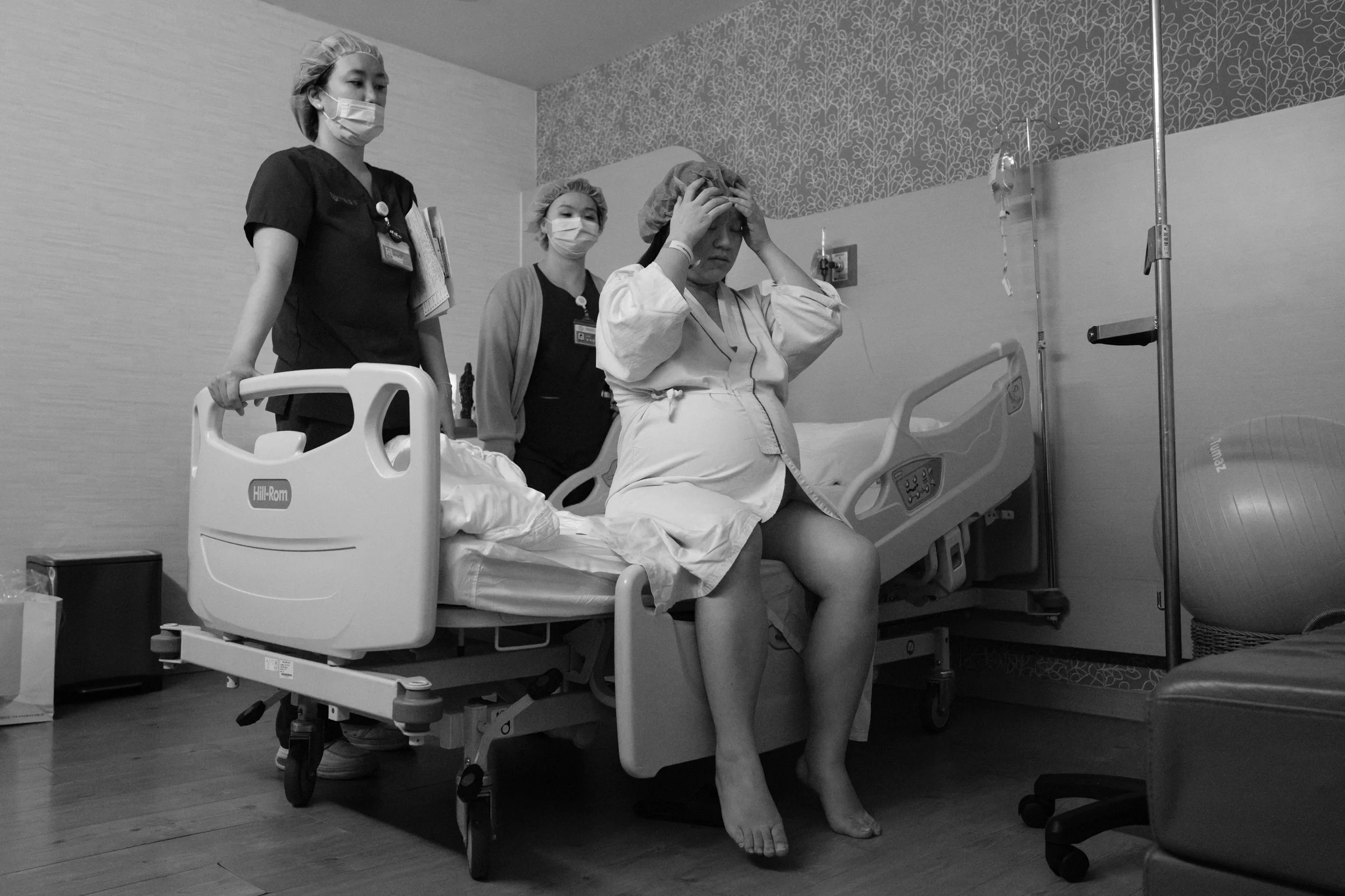 Documentary birth photography at Dianthus Taipei — mother sits on the edge of the hospital bed with head in hands as nurses prepare the room for c-section.