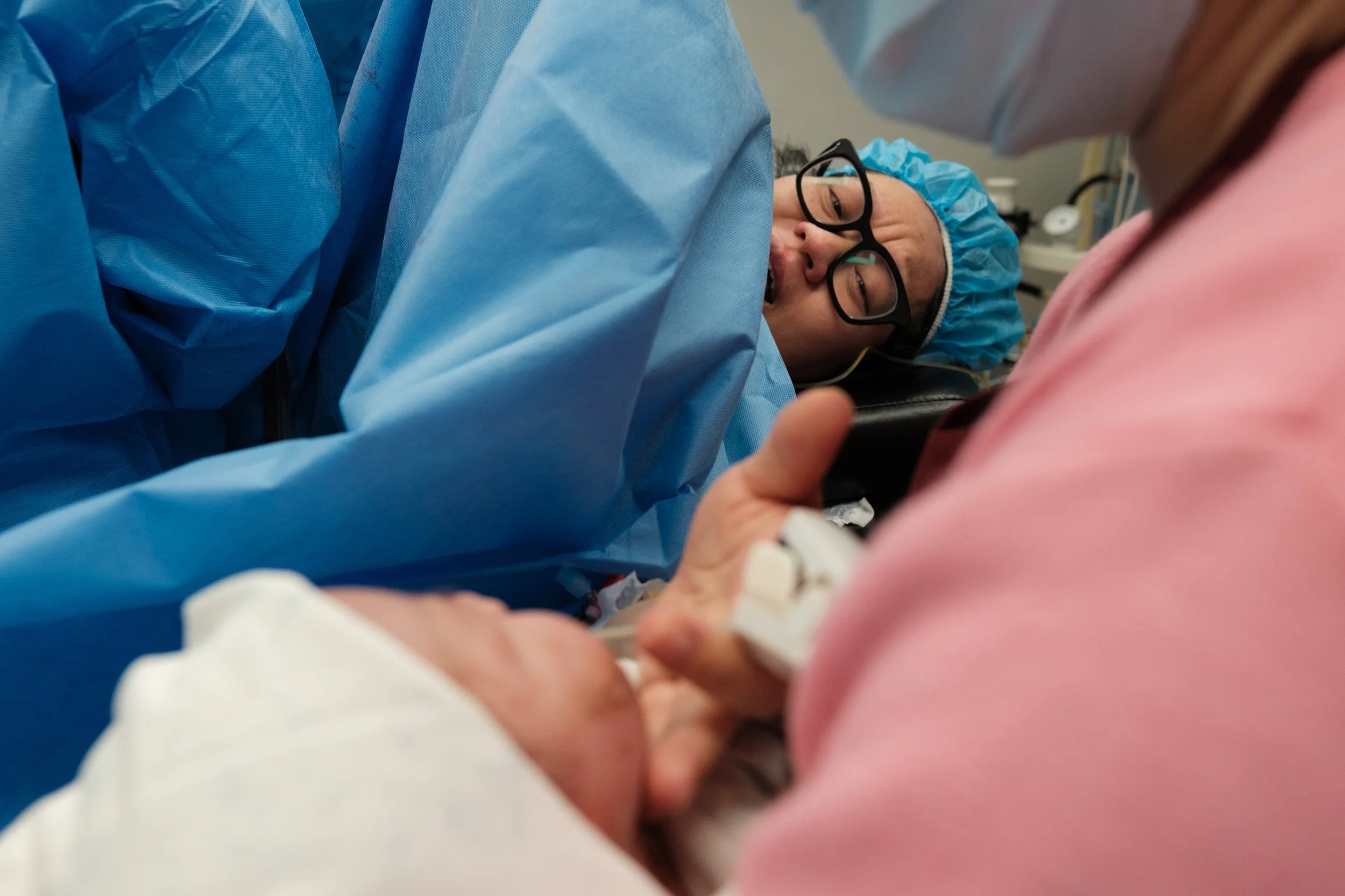 Taipei birth photography — mother's face visible as she touches her baby for the first time after a c-section surgery, partner visible in surgical gown beside her.