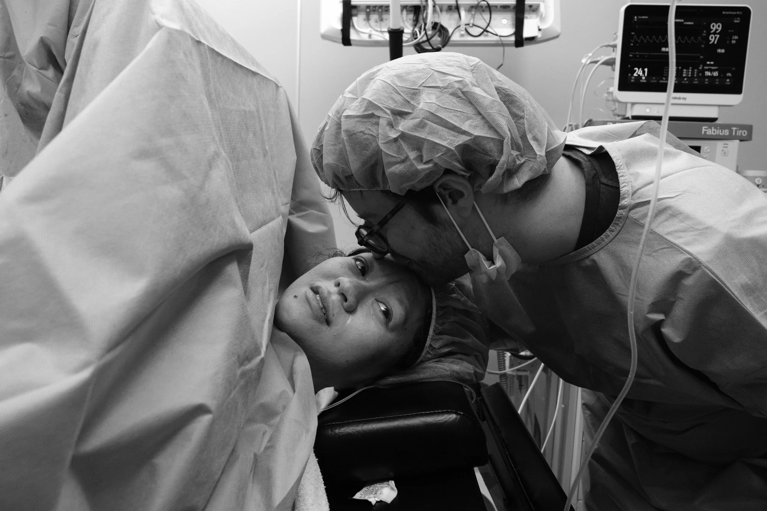 Birth photography Taipei — partner kisses mother's forehead during c-section recovery, vital signs monitor visible in background.
