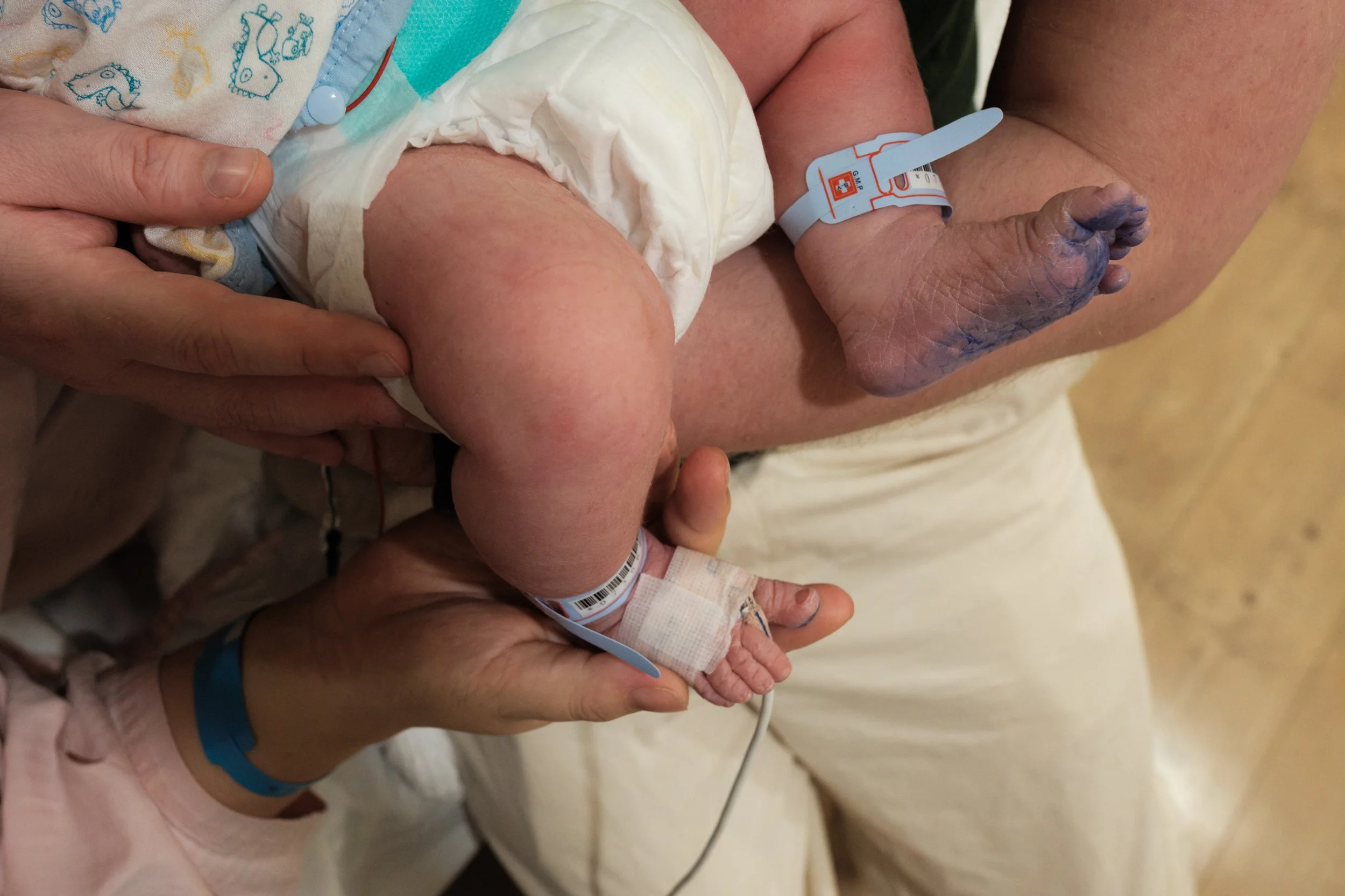 C-section birth photography Taipei — close-up of newborn's feet and hospital ID bands held in parents' hands.