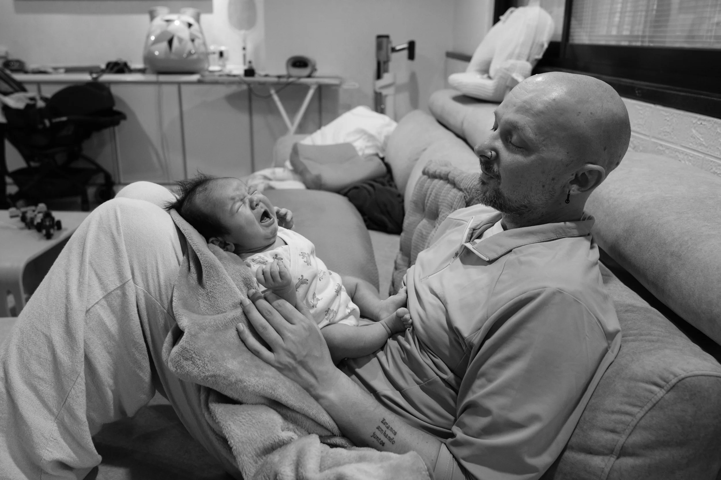 Black and white photo of a father lying on the couch with his crying newborn on his chest, candid in-home newborn session Taipei.