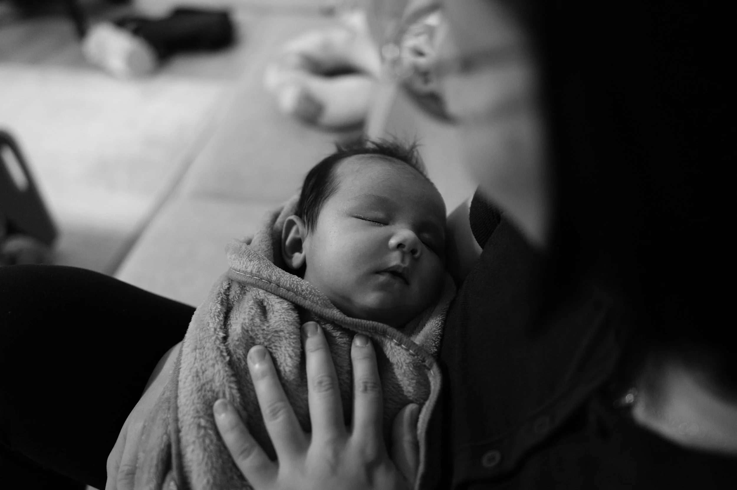 Close-up black and white photo of a sleeping newborn wrapped in a blanket with a parent's hand resting gently on the baby's chest.