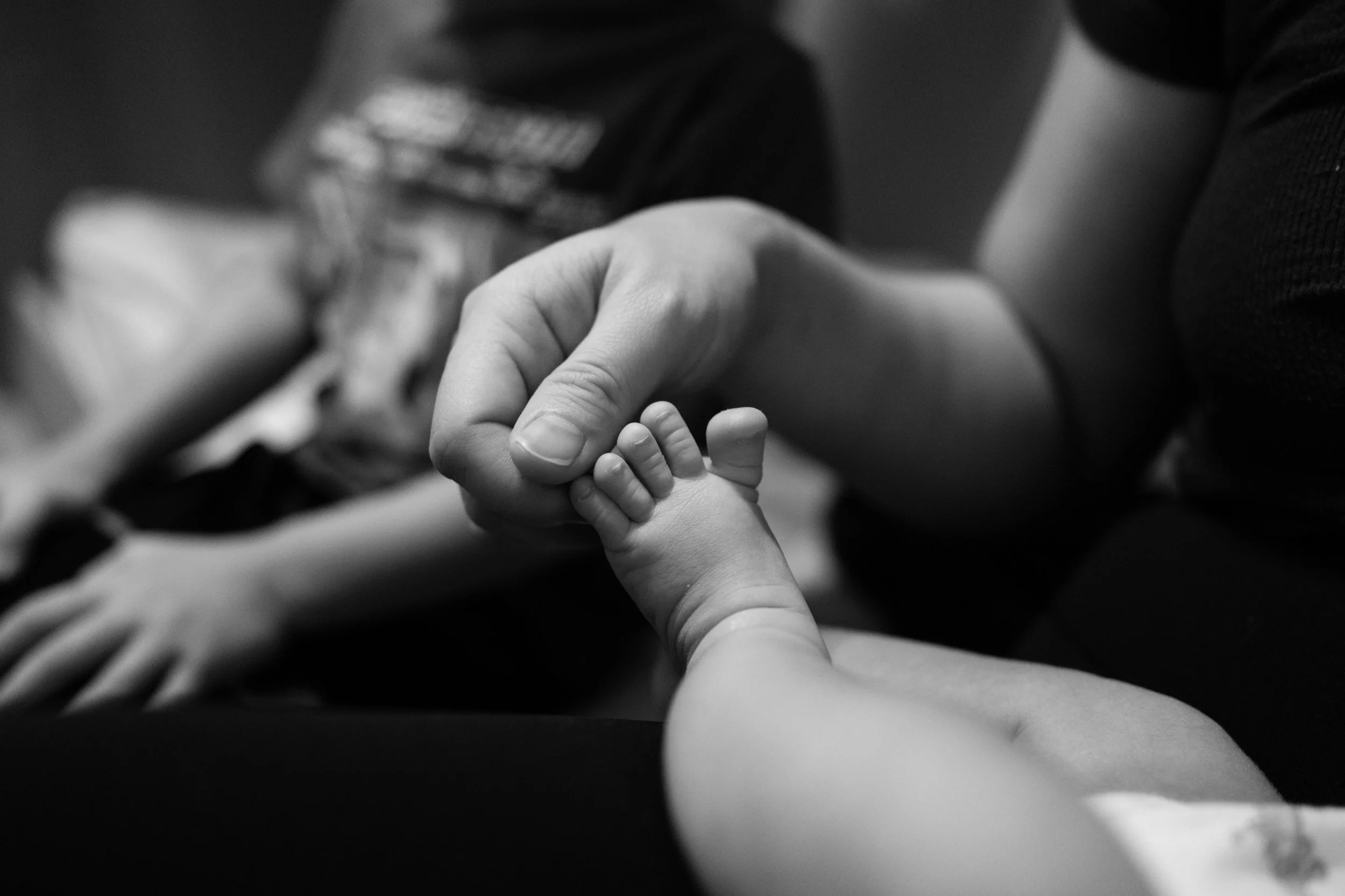 Close-up black and white photo of an adult's hand cradling a newborn's tiny foot, documentary detail shot from a Taipei newborn session.