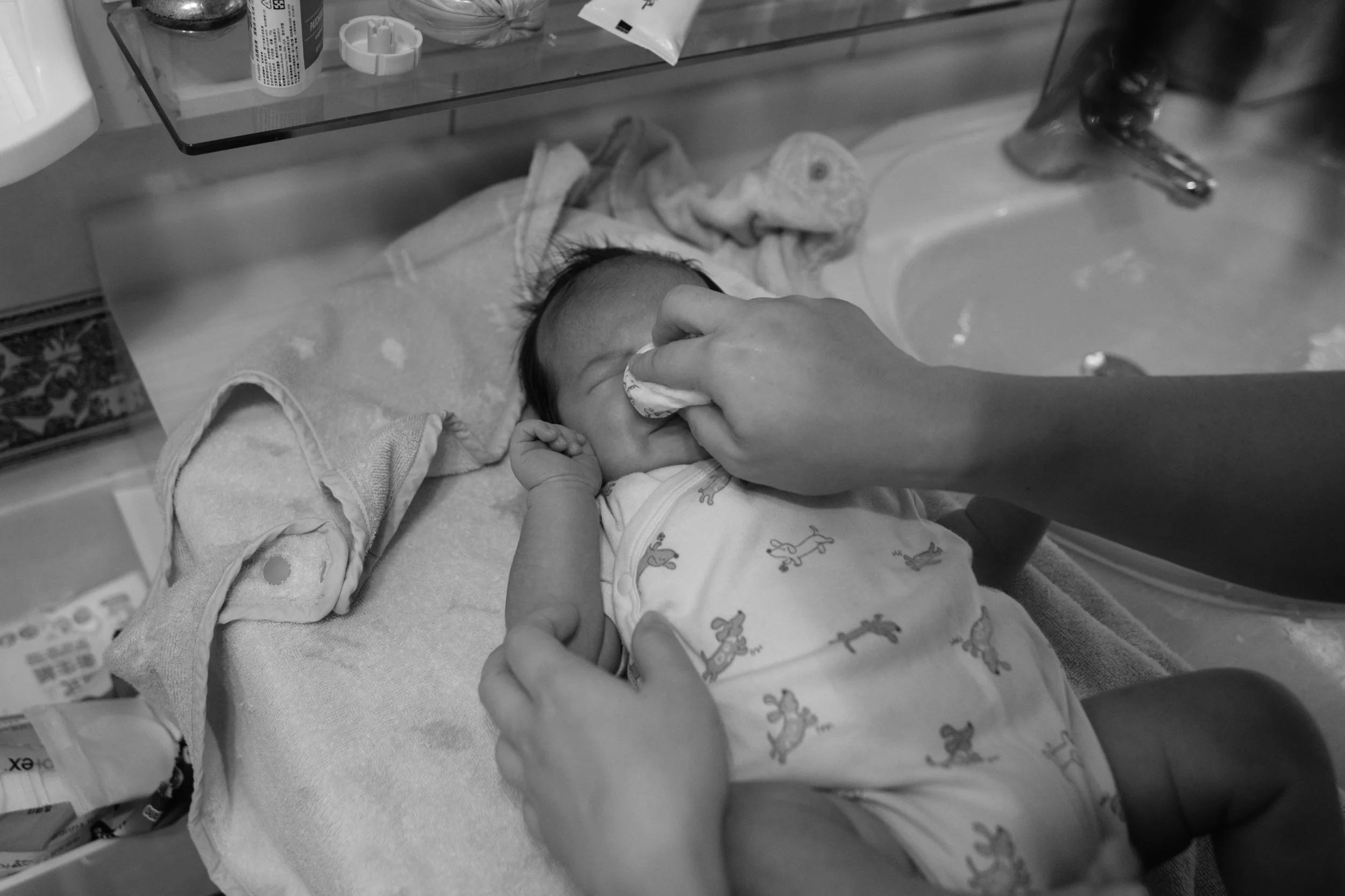 Overhead black and white photo of a parent gently wiping a newborn's face with a cotton pad during bath time in a Taipei bathroom.