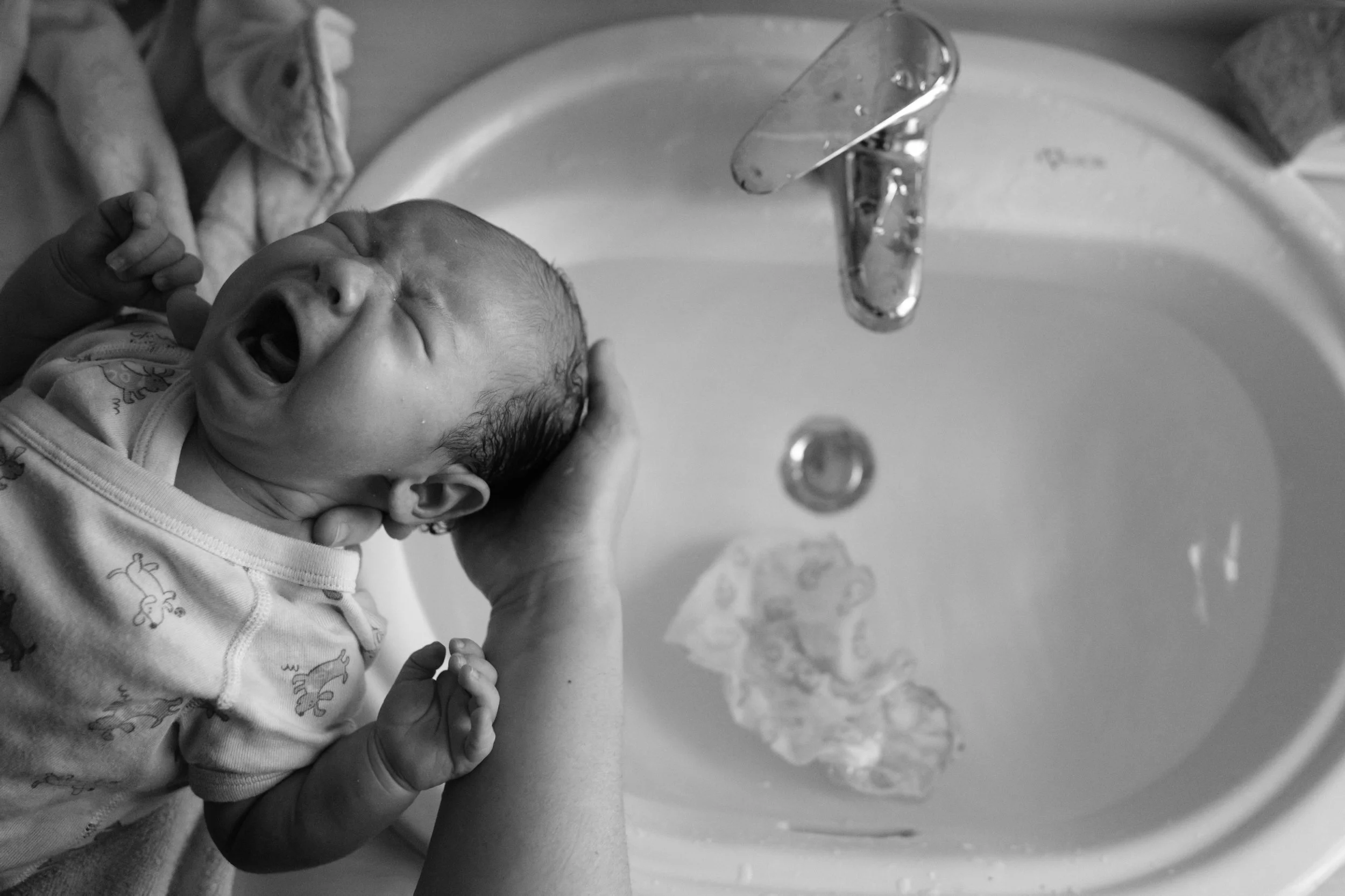Overhead black and white photo of a crying newborn being held over a bathroom sink during a home bath, documentary newborn photography.
