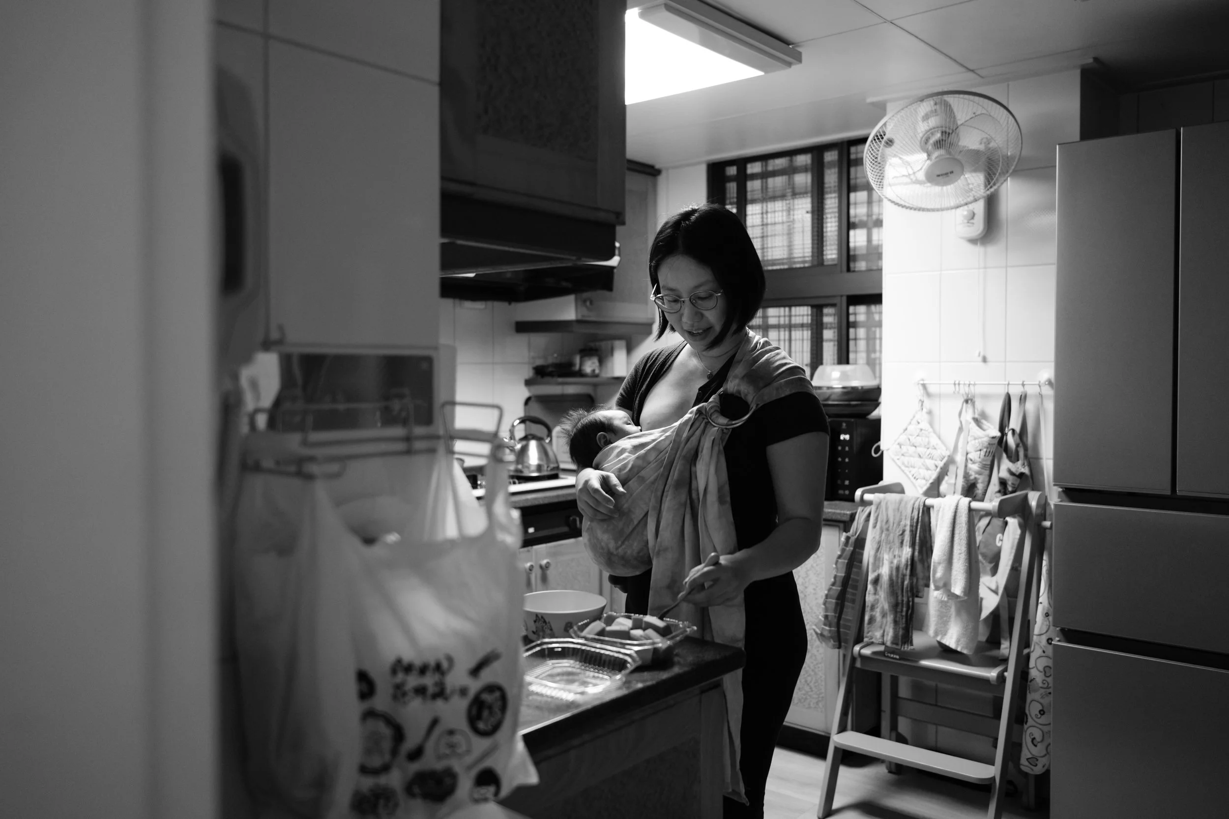 Black and white photo of a mother preparing food in the kitchen while wearing her newborn in a baby sling.