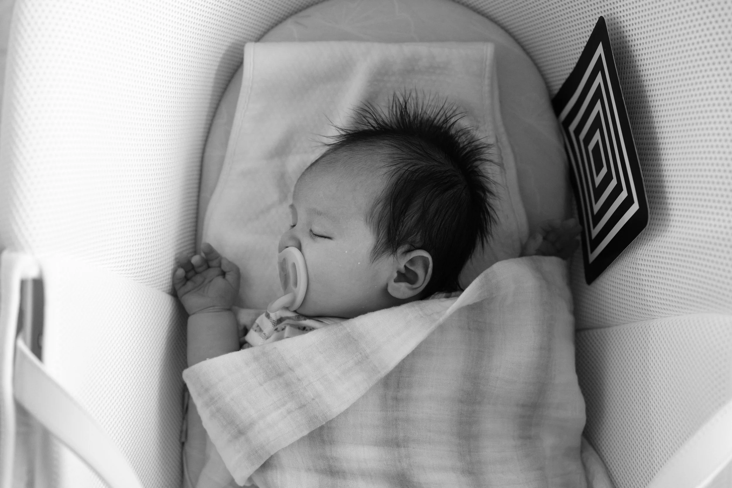 Overhead black and white photo of a sleeping newborn swaddled in a bassinet with a pacifier and a high-contrast sensory card nearby.