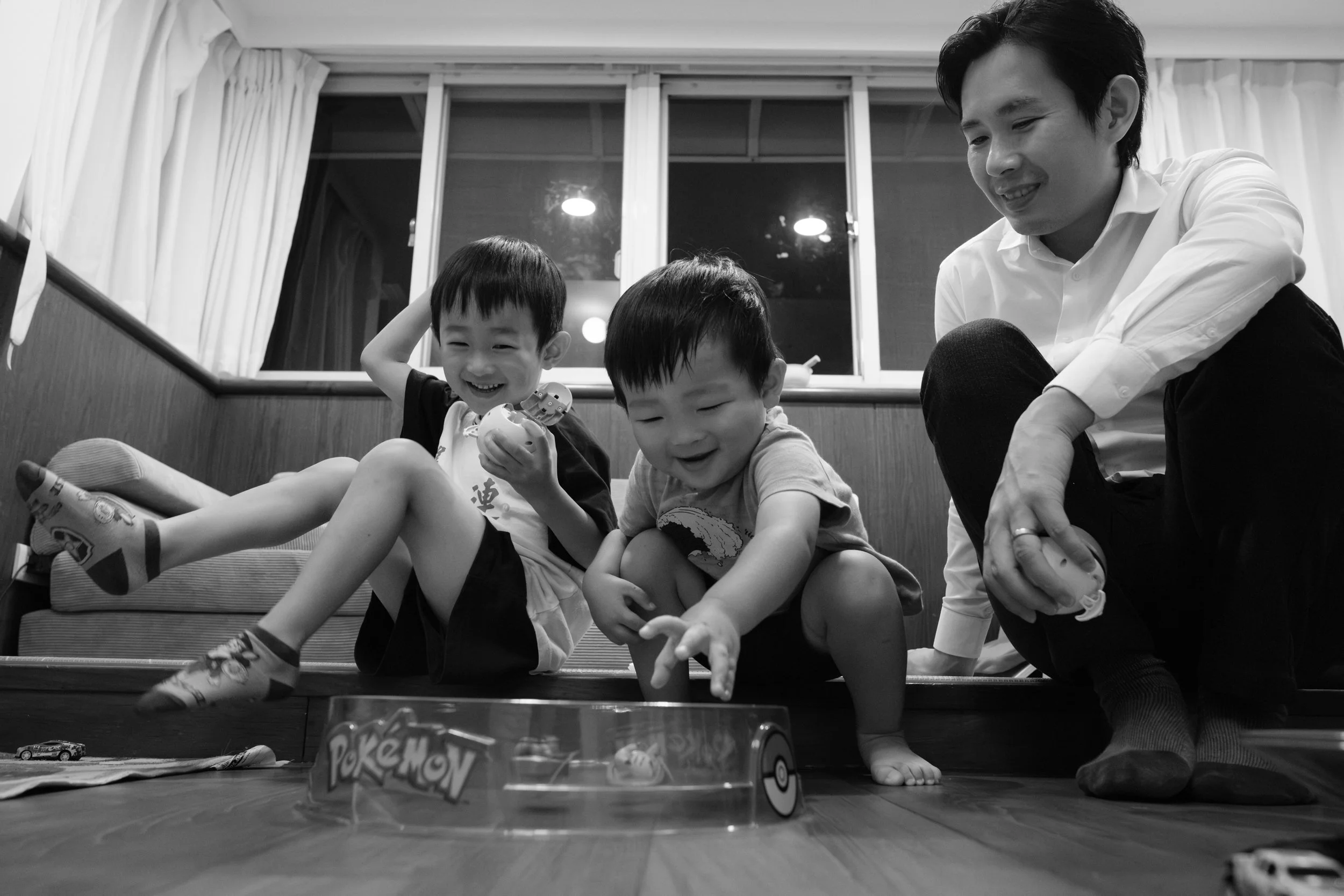 Documentary family photography in Taipei — two brothers laughing over a Pokémon toy while their father watches, part of an in-home bedtime photoshoot.
