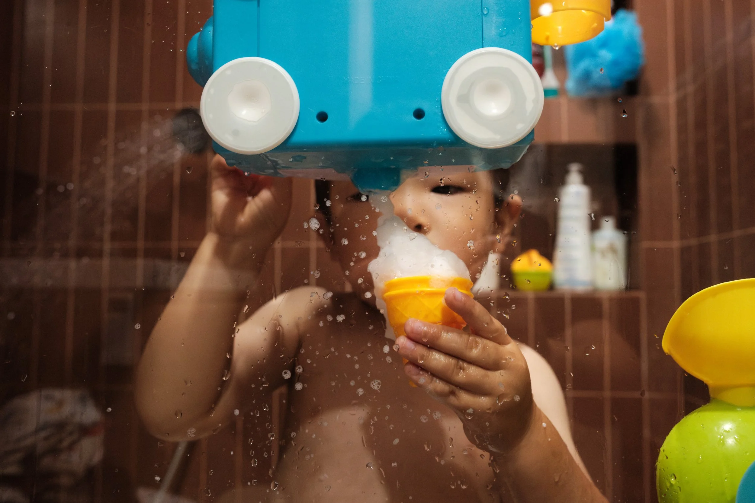 Documentary bath-time photo from a Taipei family photoshoot, child wearing a toy shower-head helmet behind a water-streaked glass door.