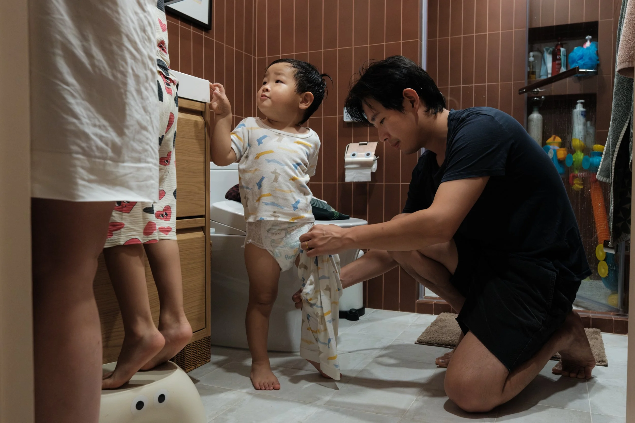 Documentary family photography in Taipei showing a father dressing his toddler in pajamas post-bath, sibling on a stool nearby.