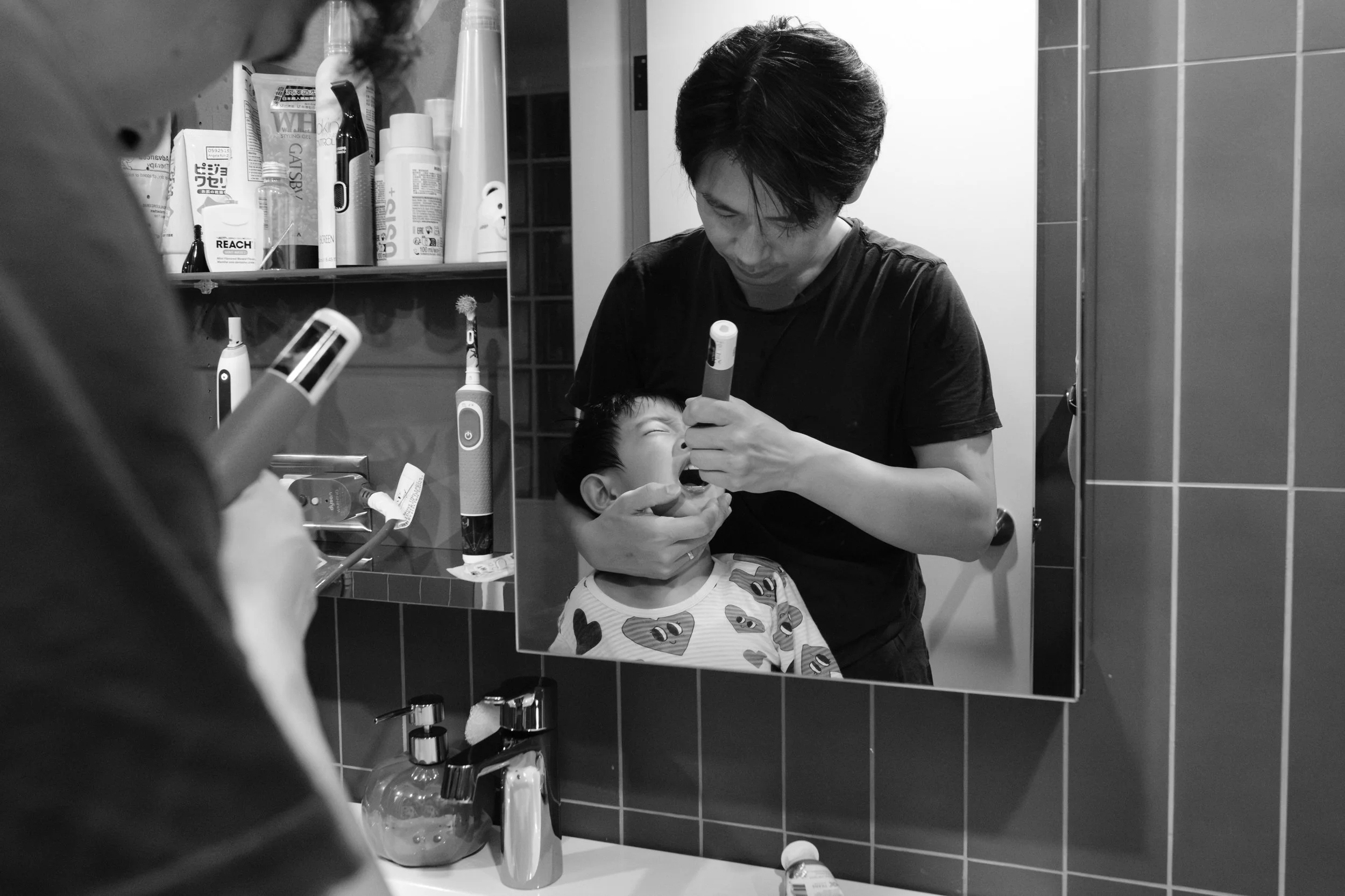 Taipei family documentary photo, mirror reflection of a father brushing his son's teeth during the nighttime routine.