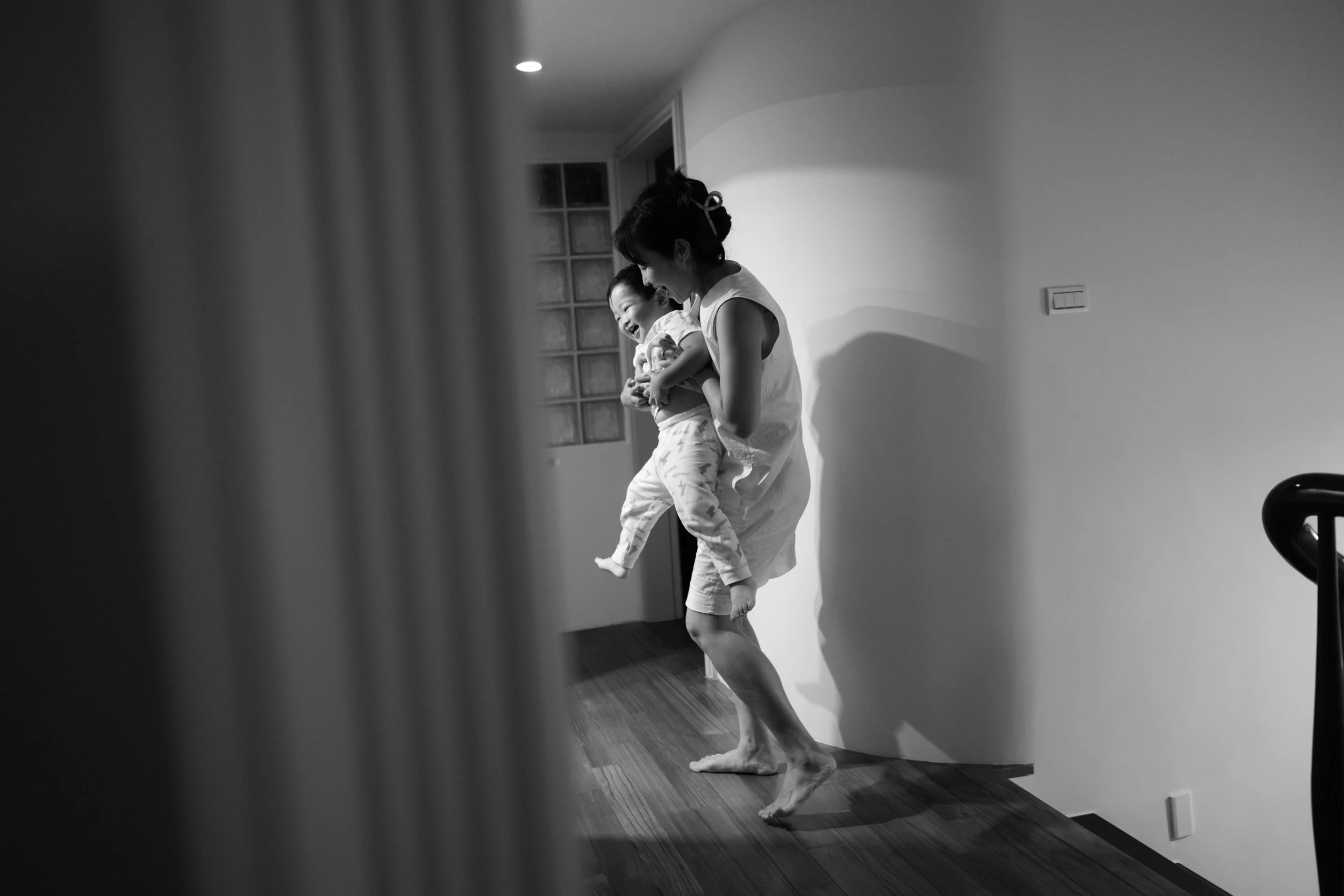 In-home family photoshoot in Taipei — mother carrying her laughing toddler down the hallway before bed.