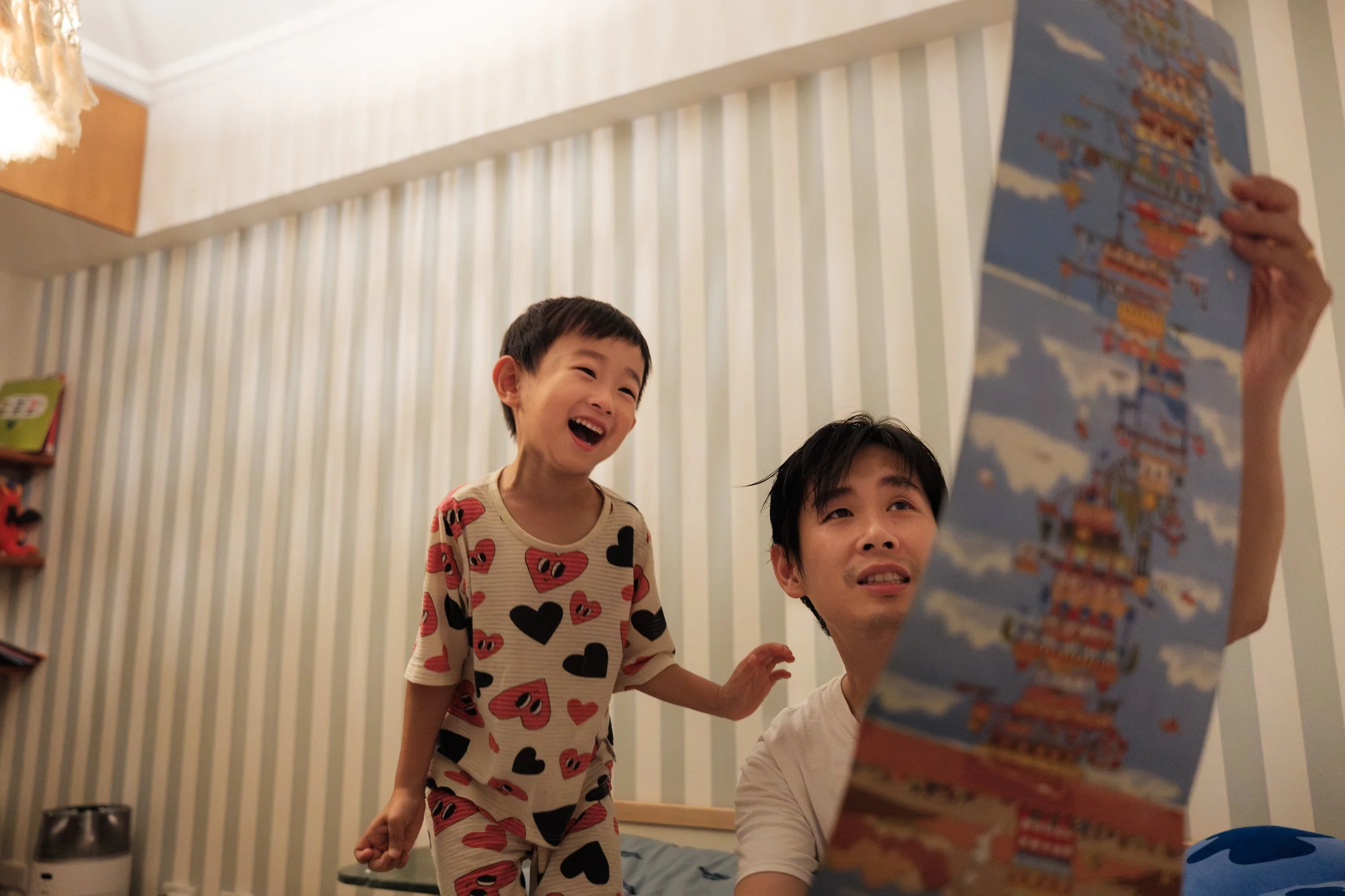Color photo from a Taipei family photoshoot showing a young boy laughing beside his father while looking at an illustrated poster.