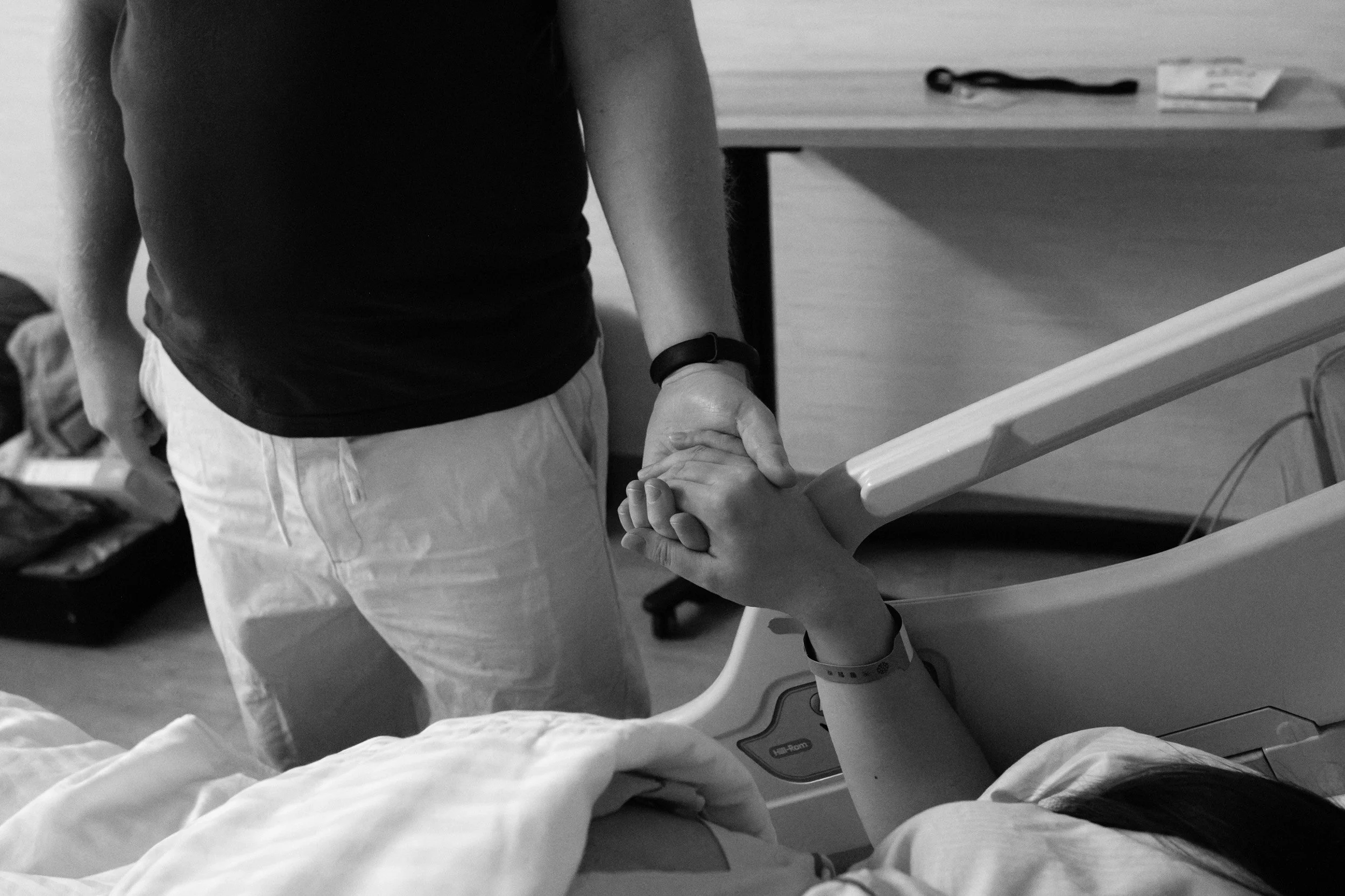 Black and white photo by a Taipei birth photographer of a husband holding his laboring wife's hand at her hospital bedside at Dianthus