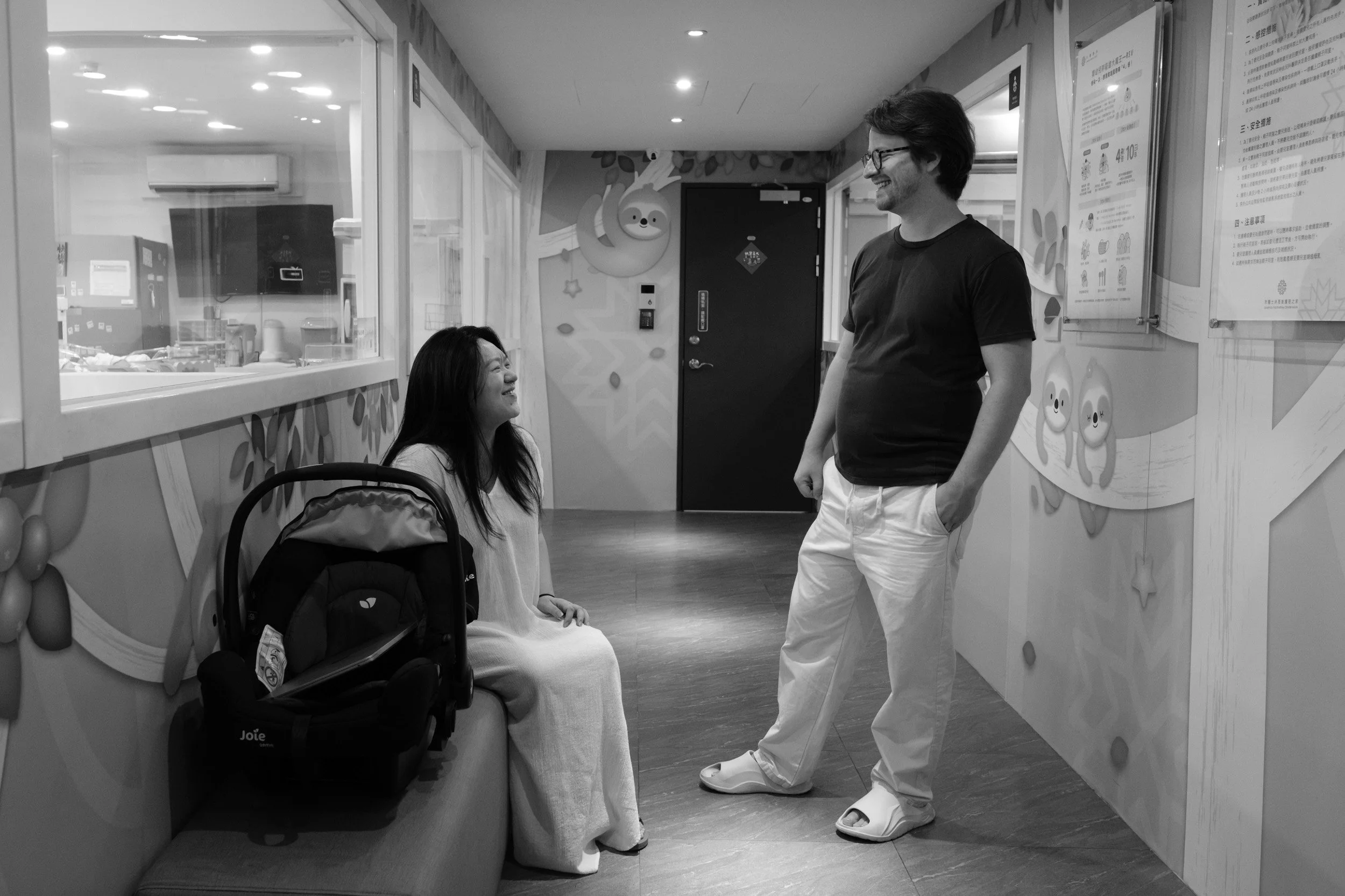 Mother sits beside her newborn's car seat in the Dianthus postpartum center hallway, smiling up at her husband before they leave, photographed by a Taipei birth photographer