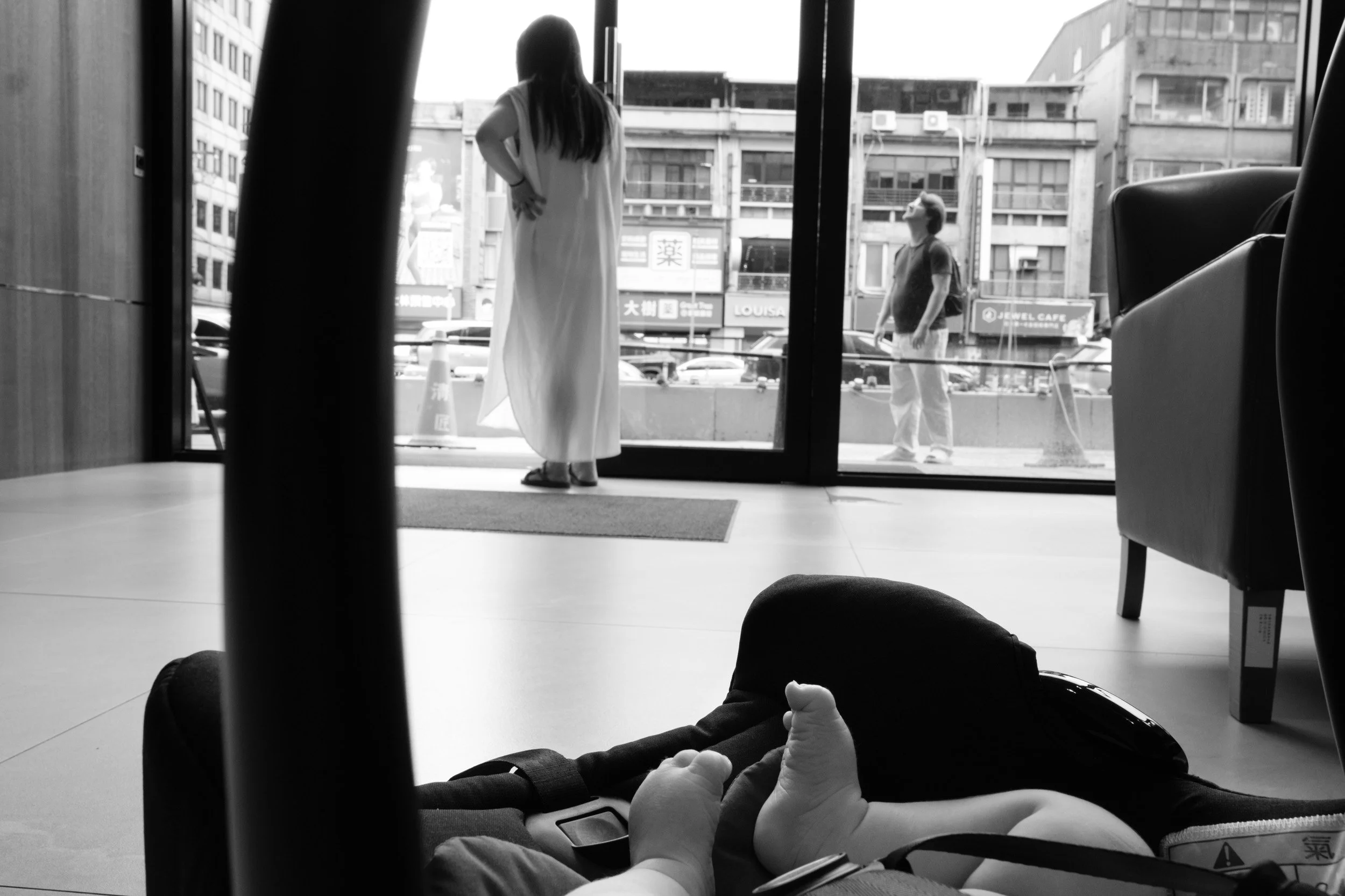 Newborn's feet in the foreground of a car seat while the parents wait at the glass doors of the postpartum center in Taipei, a newborn homecoming photographed in black and white