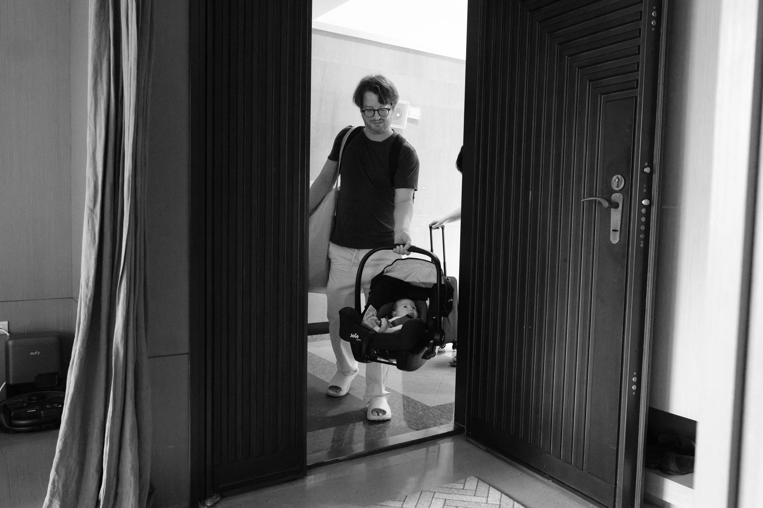 Father carries his newborn in a car seat through the front door of their home for the first time, bag on his shoulder, photographed by a Taipei birth photographer