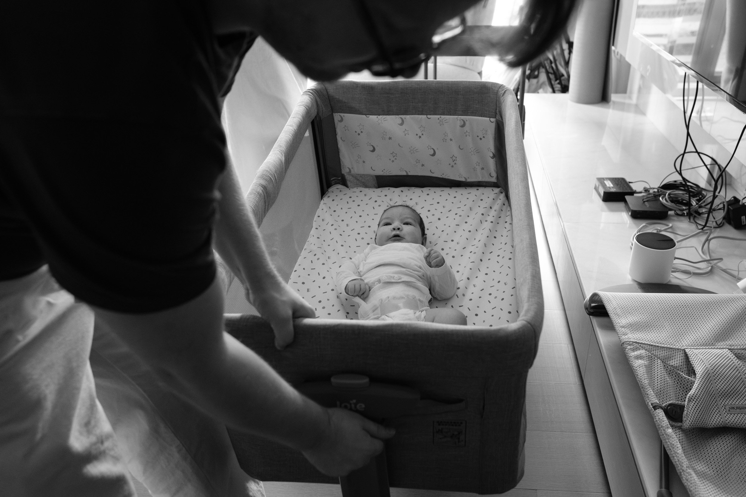 Newborn lies awake in a bedside bassinet as a parent leans over it, a homecoming session photographed in Taipei