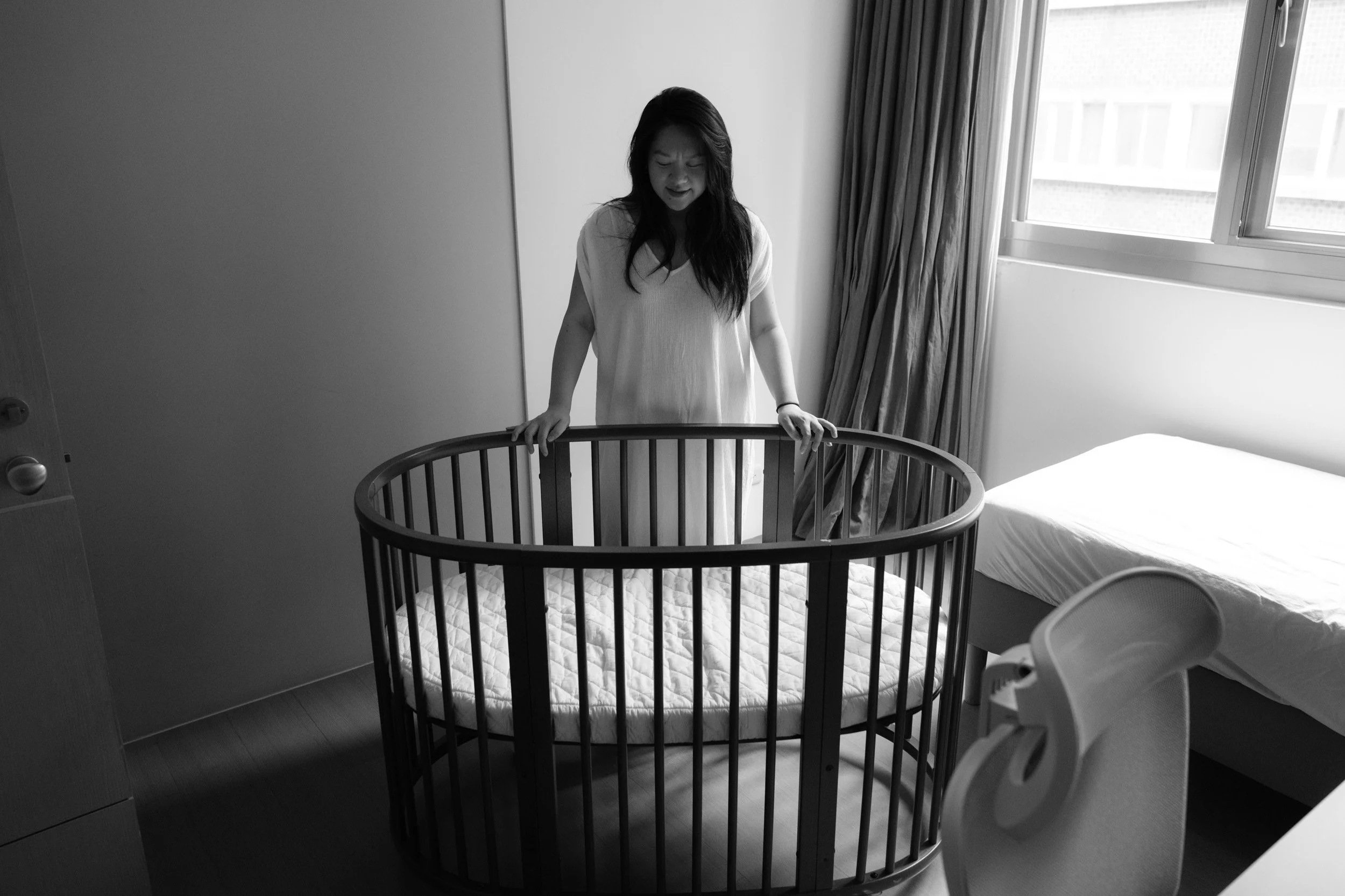 Mother in a white dress stands at an empty oval wooden crib, smiling as she looks down, in her Taipei home before the newborn's first night