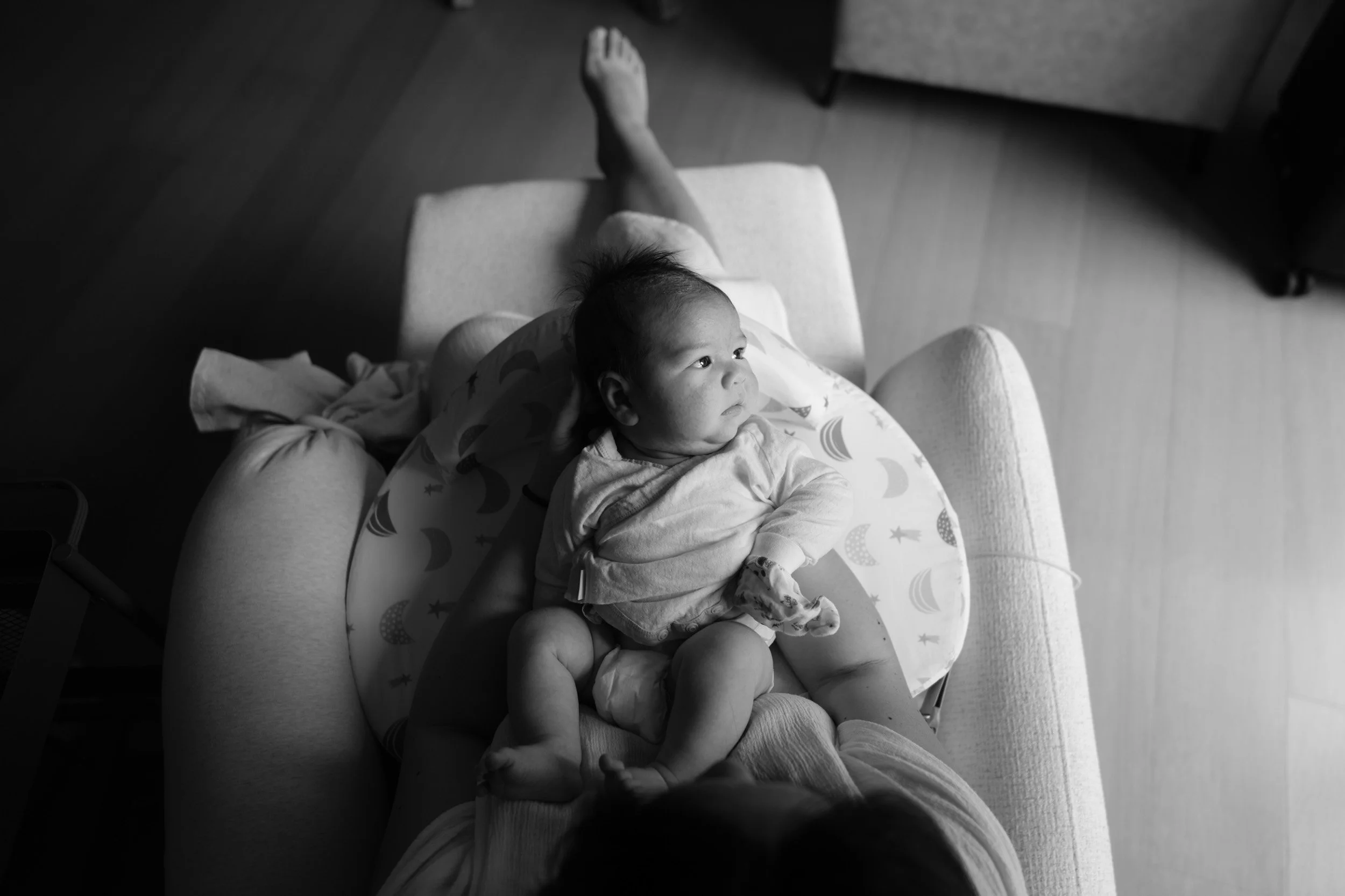 Overhead view of a newborn seated on a moon-and-stars nursing pillow in his mother's lap, gazing off to the side, during a newborn homecoming session in Taipei