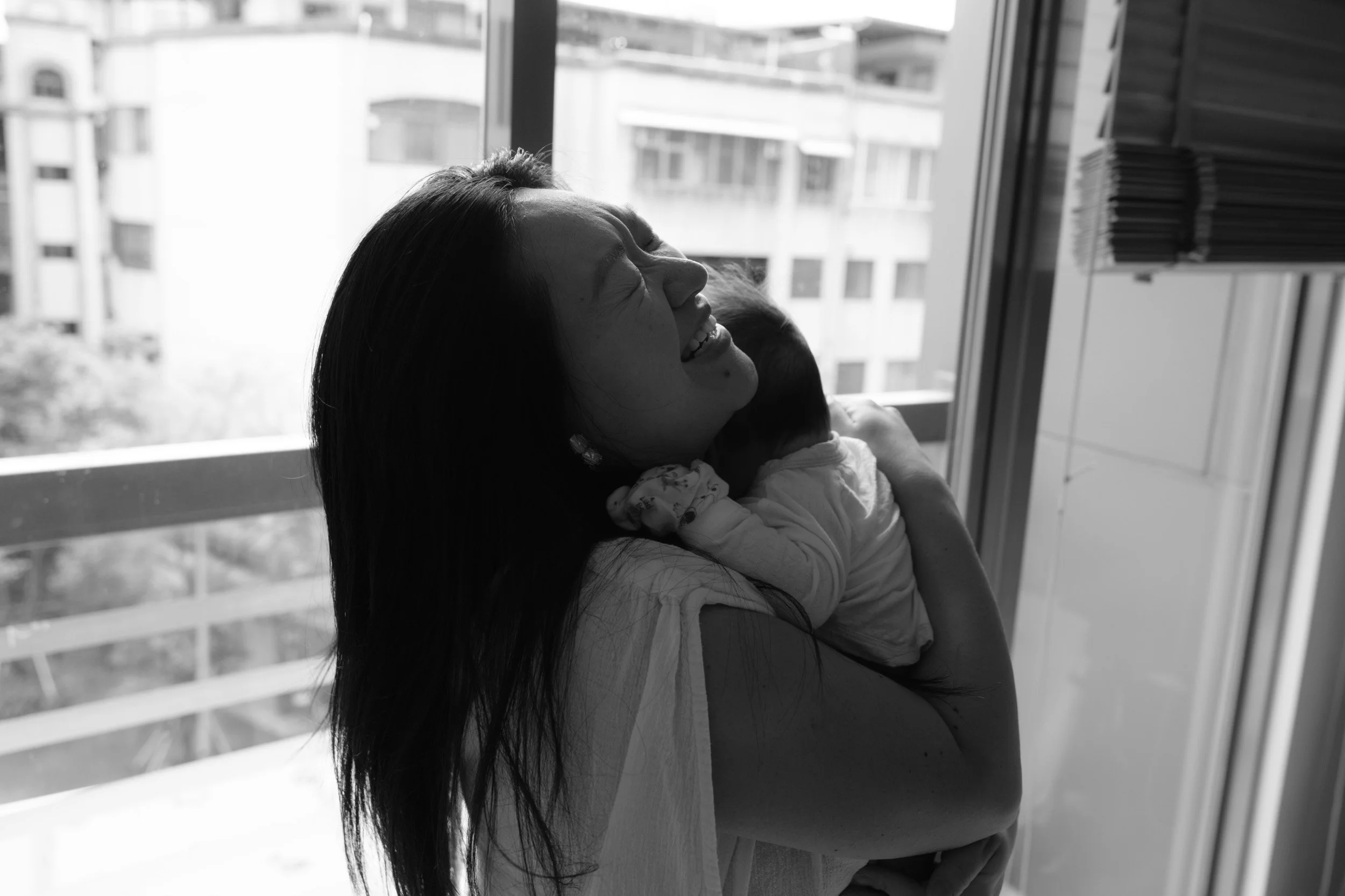Laughing mother with her eyes closed holds her newborn against her shoulder by a bright window, a candid moment from a Taipei homecoming session