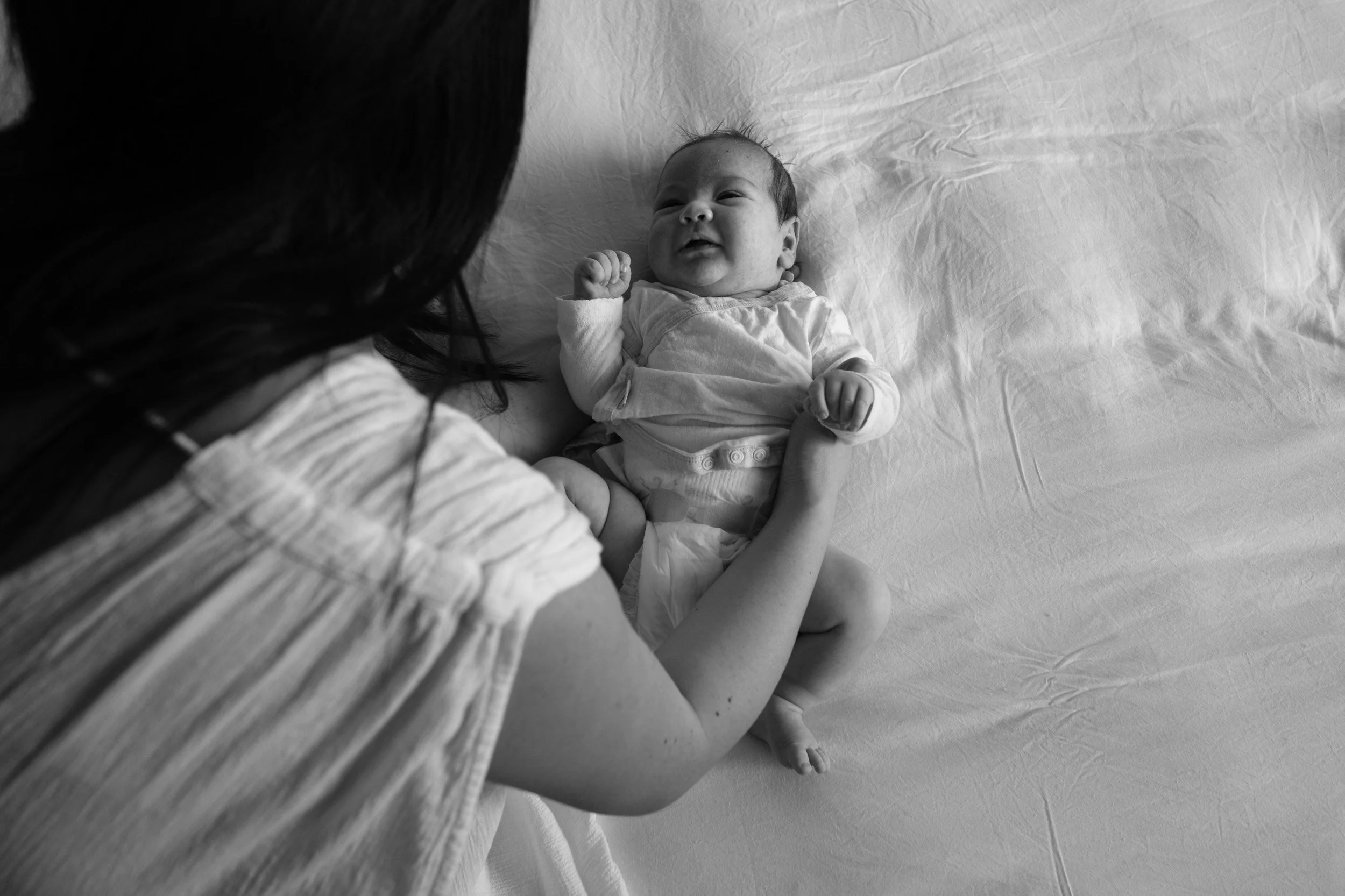 Smiling newborn lies on a white bed looking up at his mother, who leans in close, during a newborn homecoming session in Taipei