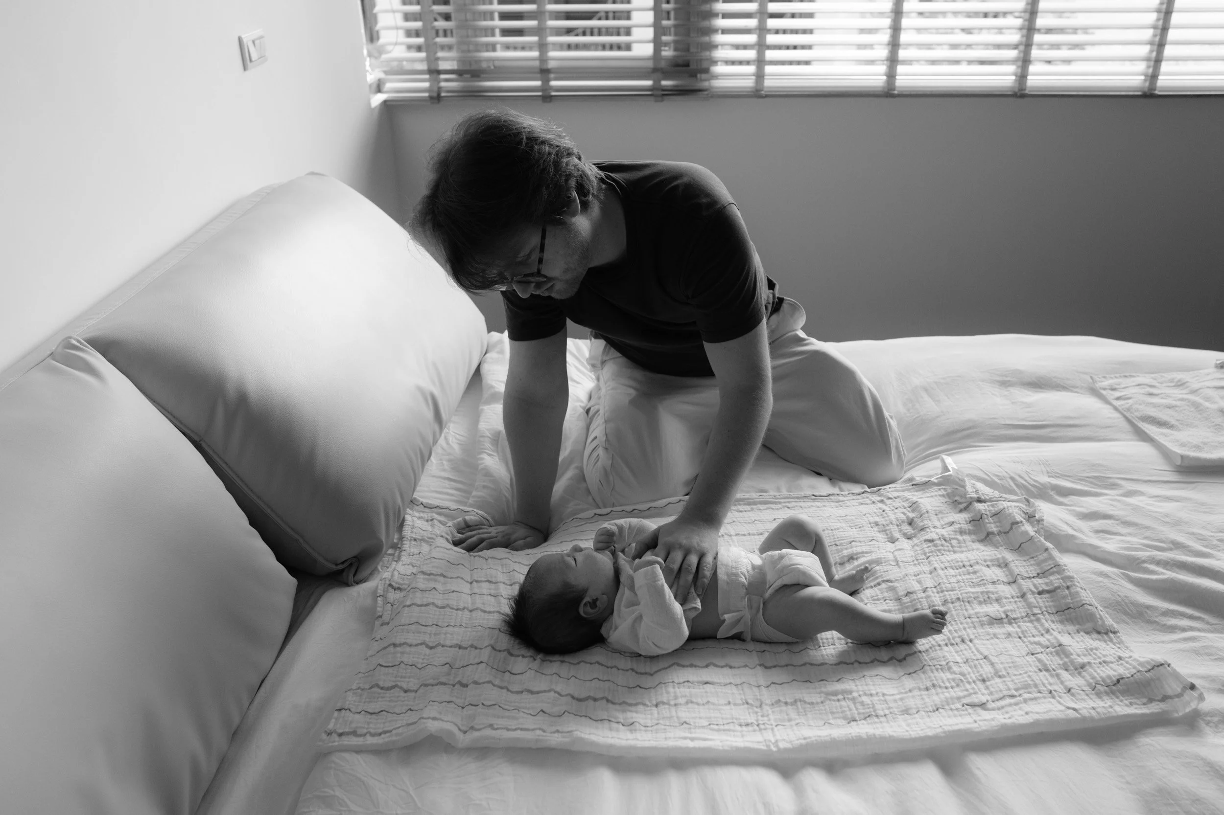 Father kneels on the bed with one hand on his newborn, who lies on a muslin blanket, photographed by a Taipei birth photographer during a homecoming session