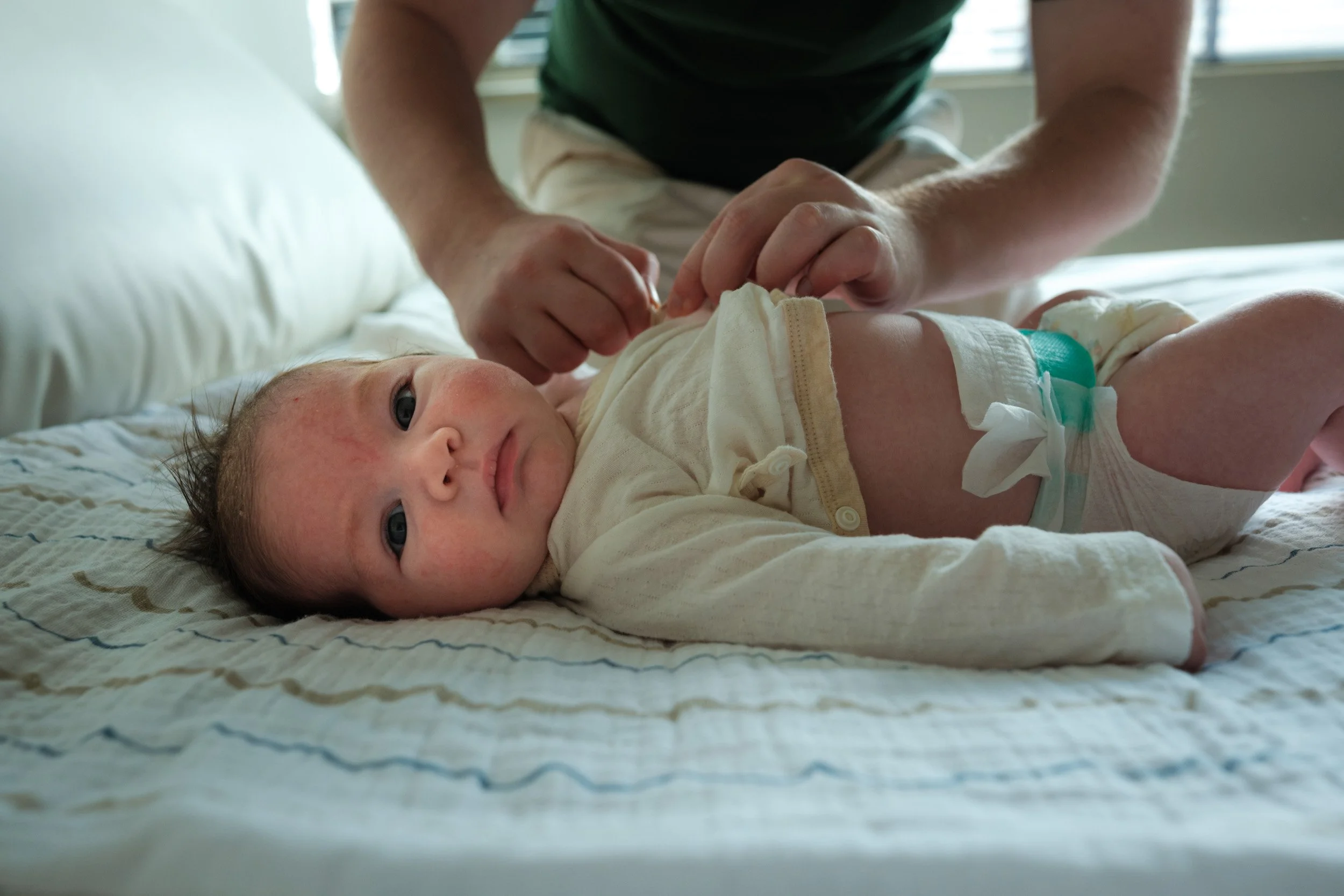 Color photo of a father's hands fastening his newborn's kimono-style top during a diaper change, while the baby looks straight at the camera, at home in Taipei