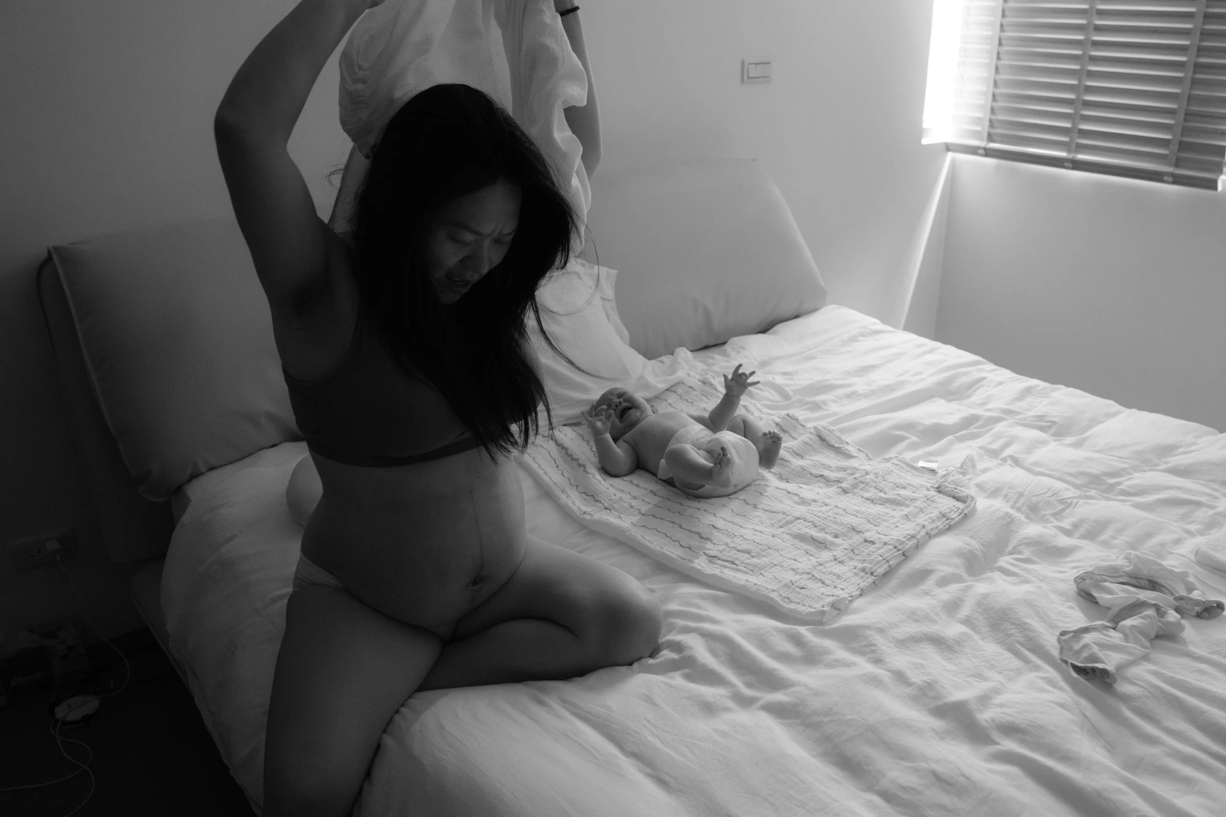 Mother pulls a shirt over her head beside her newborn, who cries on a muslin blanket on the bed, a candid postpartum moment at home in Taipei