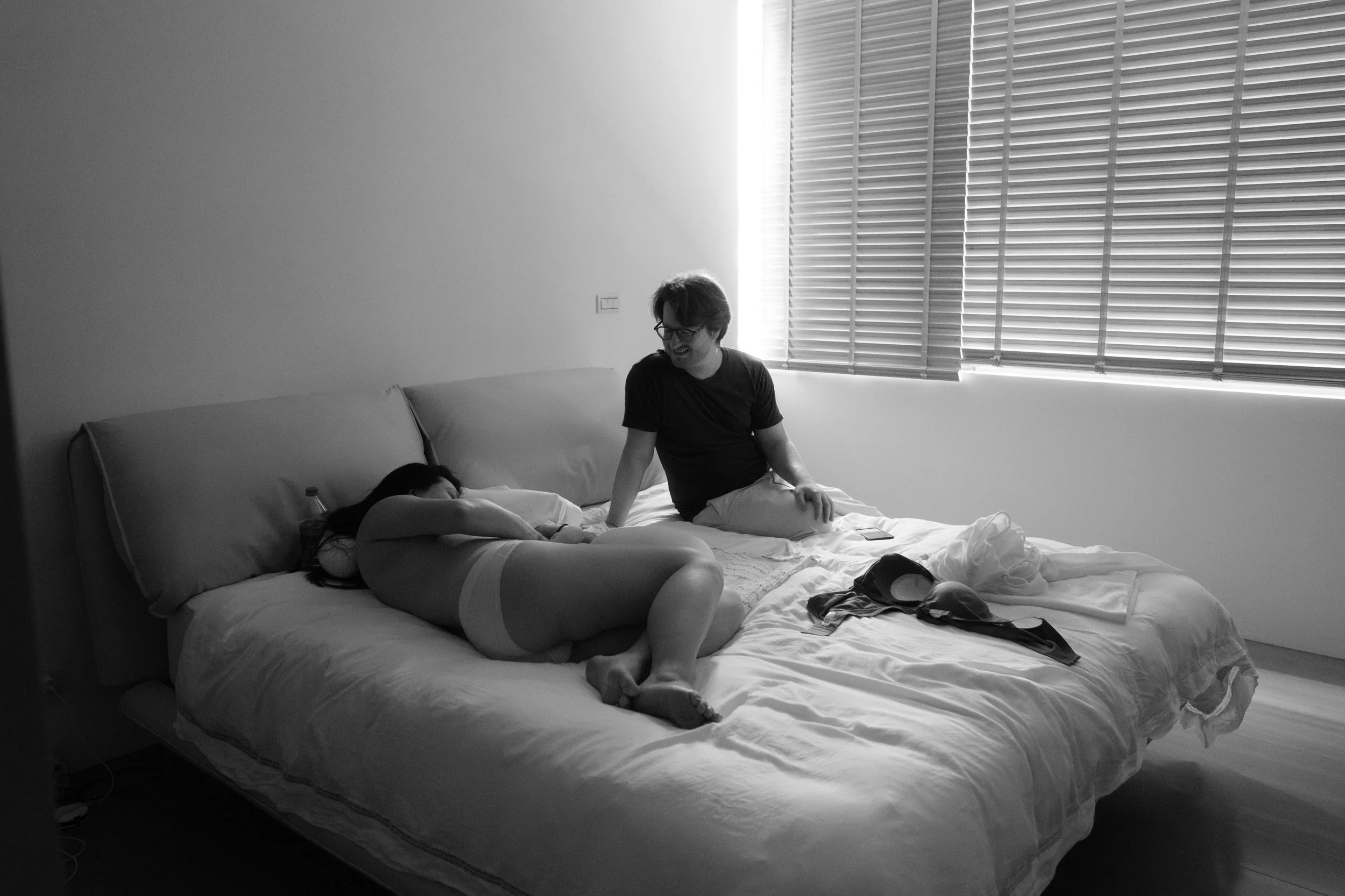 Father sits on the bed smiling at his exhausted wife, who lies curled on her side with her nursing bra on the bed beside her, at home in Taipei after the birth