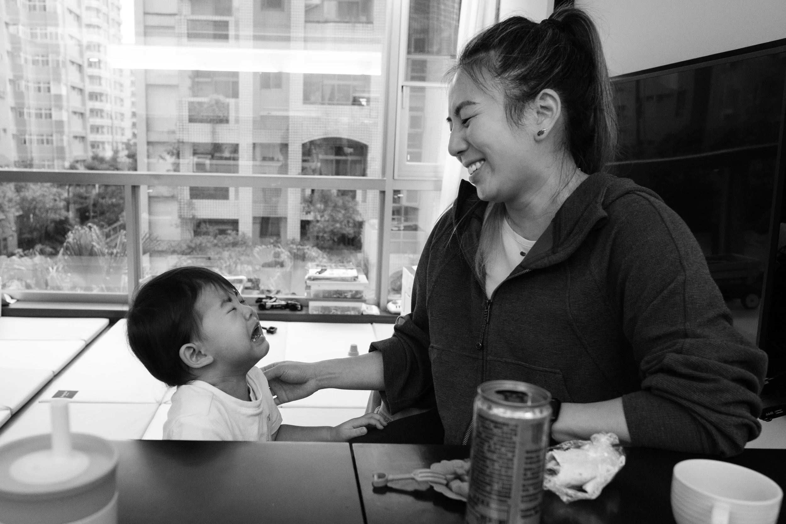 Black and white documentary family photography in Taipei: a toddler cries up at a woman who laughs at the kitchen counter