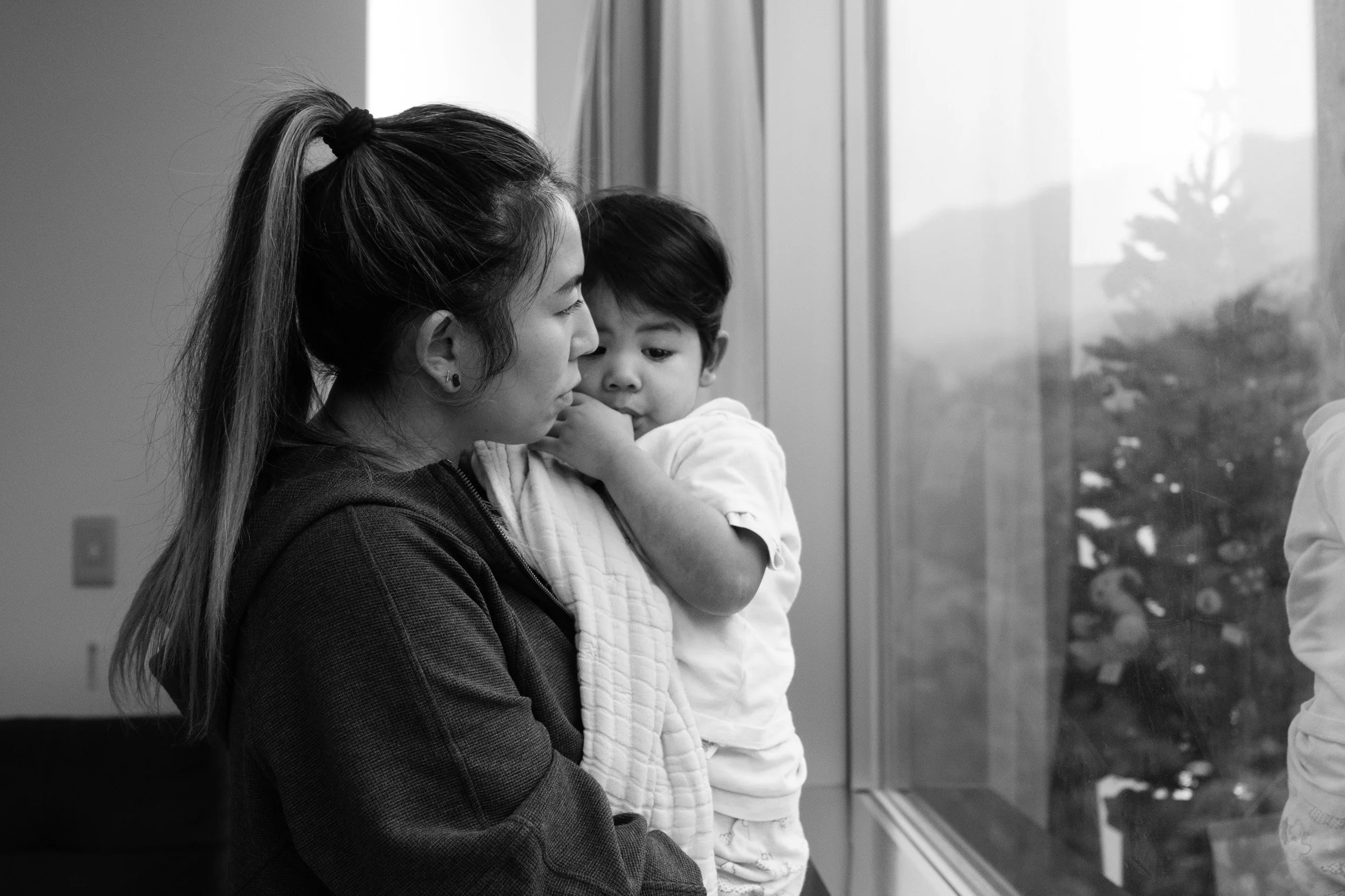 Black and white photo of a woman holding a toddler at a window, a Christmas tree reflected in the glass, by a Taipei family photographer
