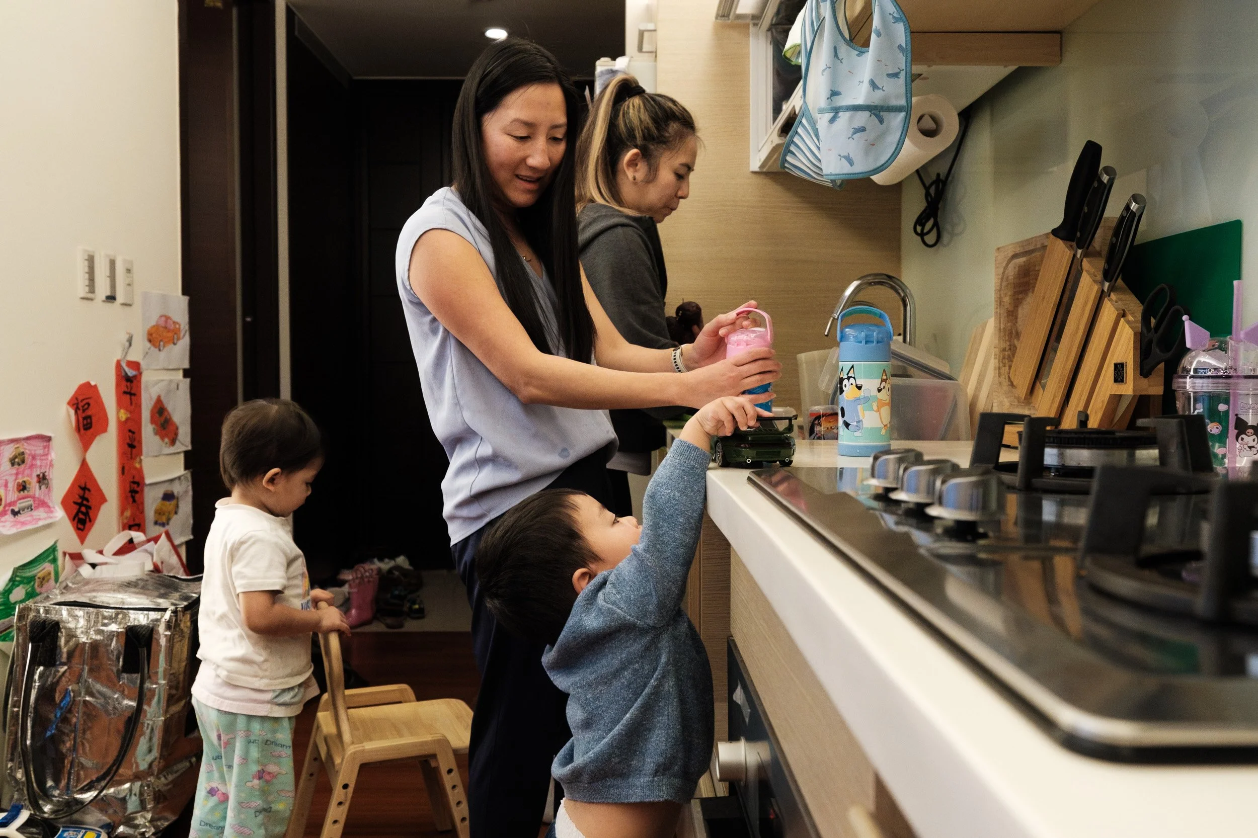 A woman fills a water bottle in a Taipei home kitchen while a toddler reaches up to the counter and another stands behind, documentary family photographer Taiwan