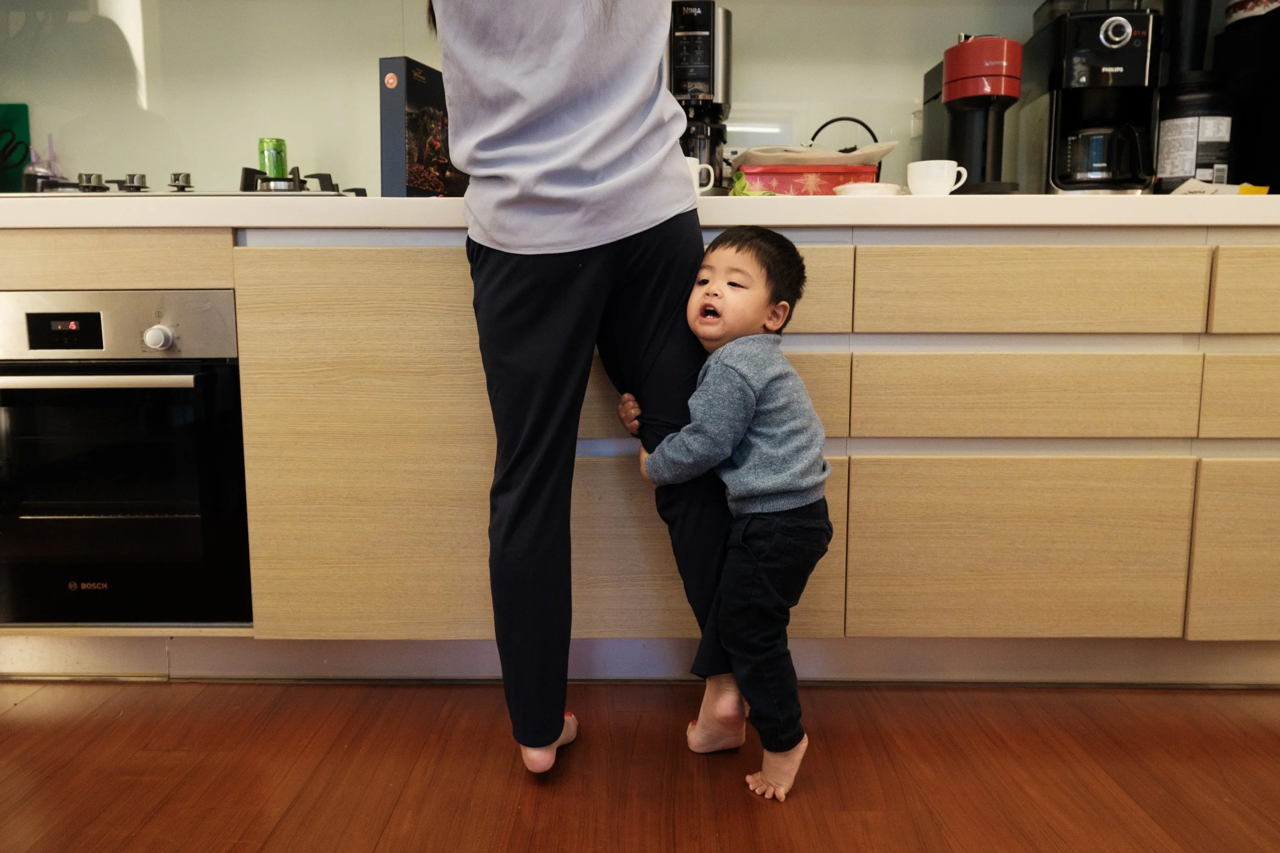A toddler clings to a woman's leg at the kitchen counter, candid Taipei family photography at home