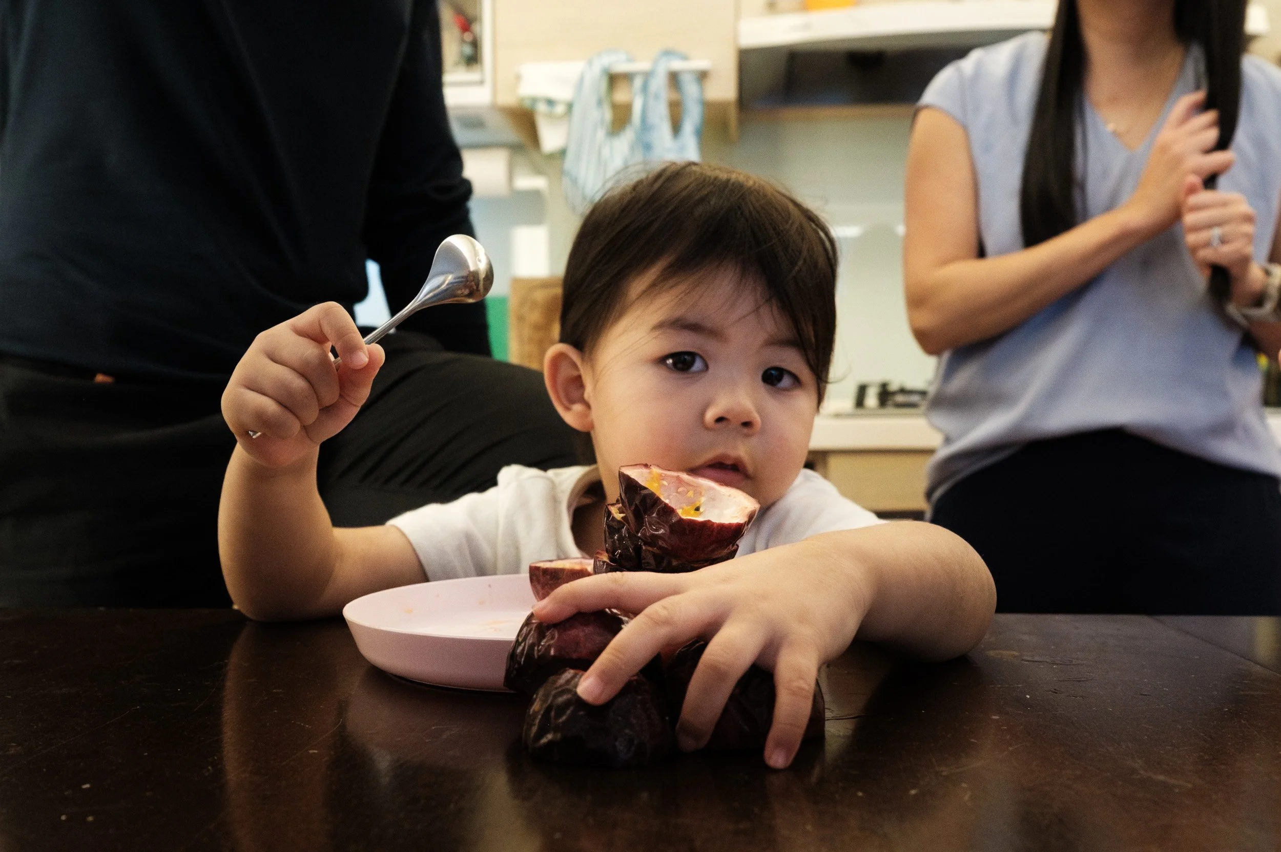 A toddler with a spoon and a stack of passion fruit at a kitchen table, unposed Taipei family photographer